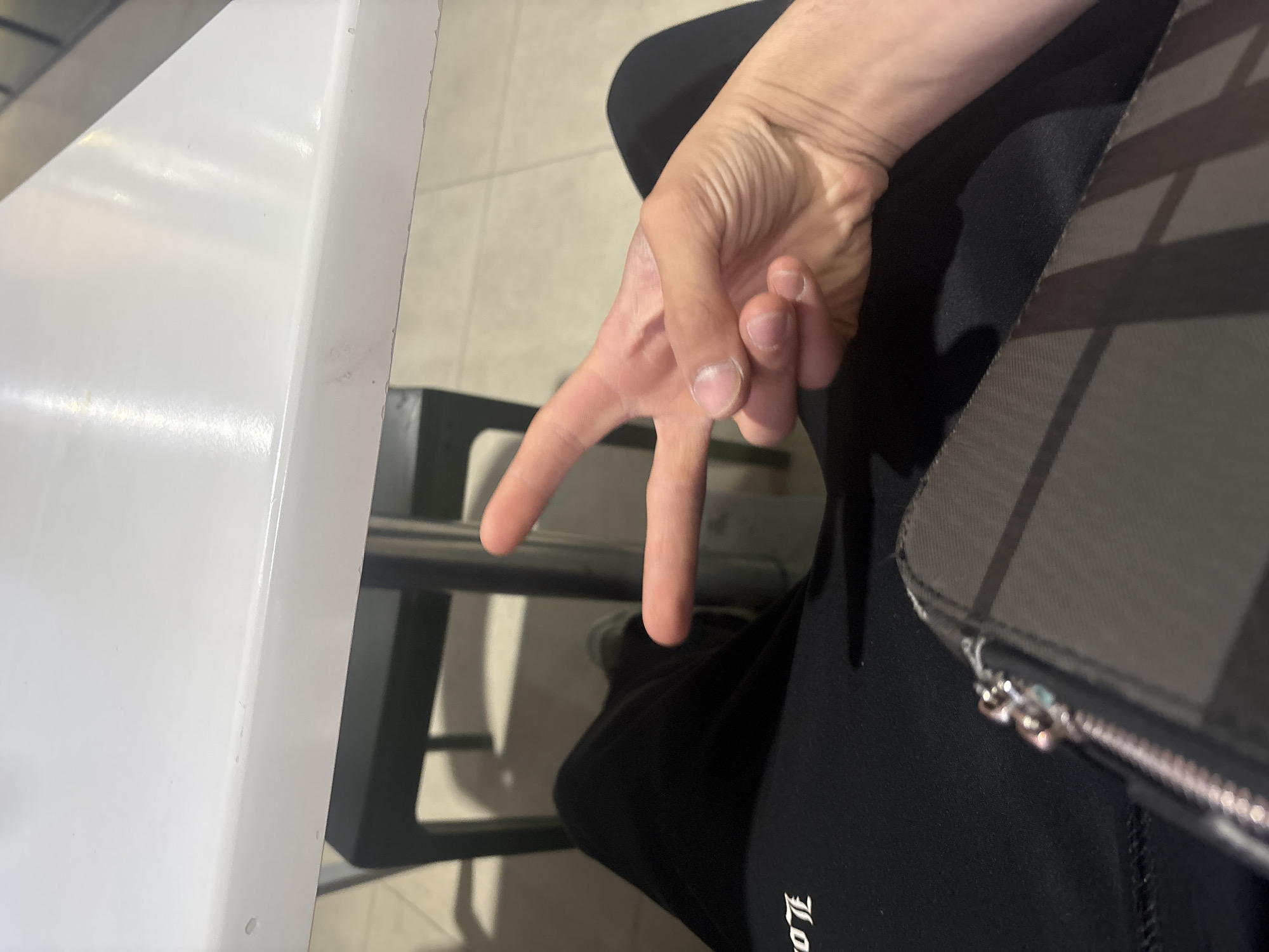 This image, taken in Yerevan, Armenia, captures a close-up, tilted view of a person's right hand making a peace or victory sign. The index and middle fingers are extended upwards in a V-shape, while the other fingers are curled into the palm. The hand has a light skin tone with visible veins and skin texture.
The person appears to be seated indoors. A clean, white table surface occupies the left side of the frame, with a dark metal chair frame partially visible beneath it. The individual is wearing dark clothing, possibly sweatpants or track pants, on which a white, partially visible gothic-style letter or logo, possibly "LO" or "JL," is embroidered. To the right of the hand, a dark, structured bag or purse with a textured surface and a metallic zipper is visible. The floor in the background is tiled with light-colored squares. The lighting is bright and even, consistent with an indoor setting during the day or evening. No specific activity beyond the hand gesture is depicted, which typically conveys positivity or acknowledgement.