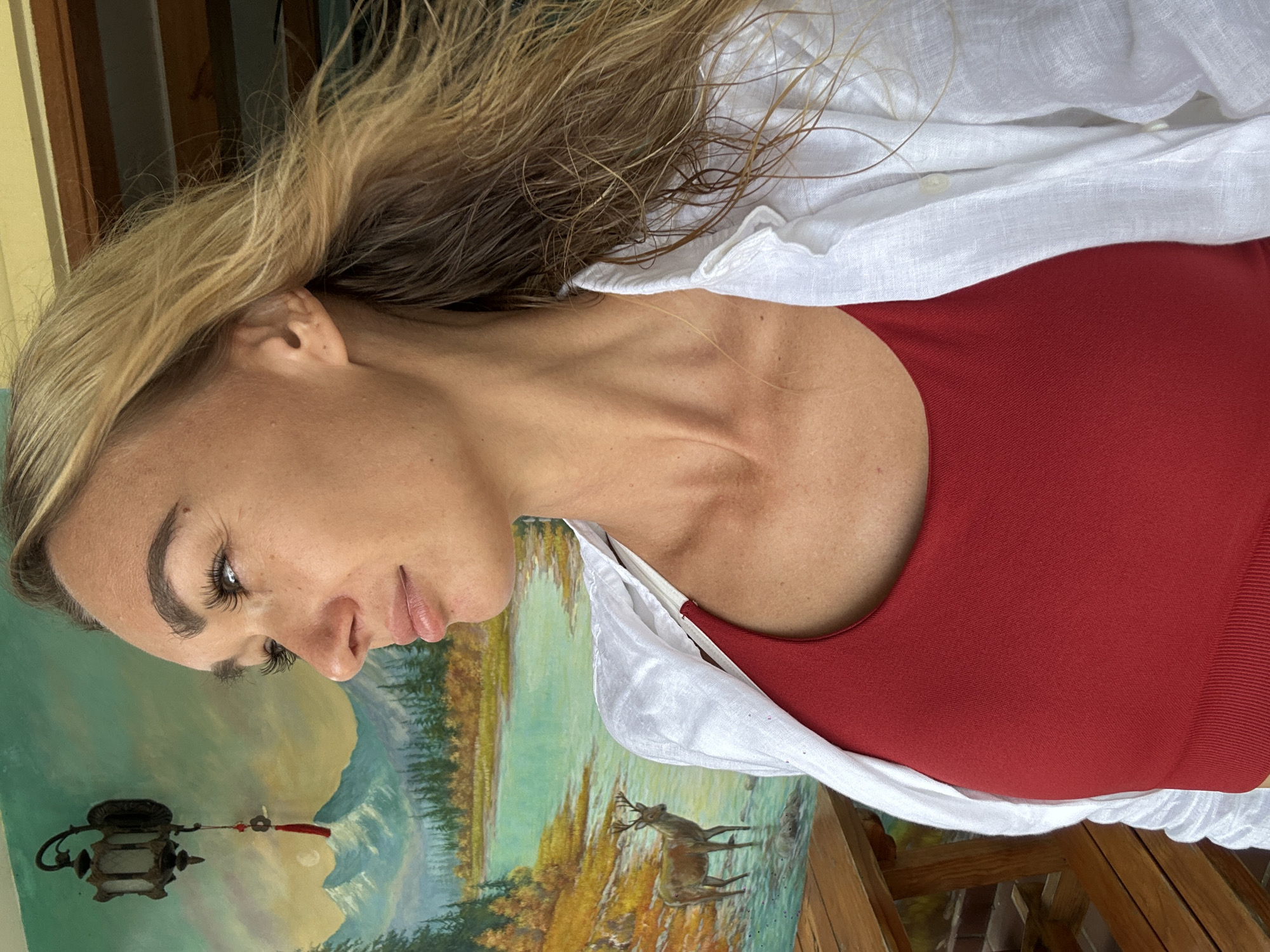 A woman with blonde hair is captured in a close-up, profile shot, looking to the left. She is wearing a red tank top and an open white linen shirt. The background features a mural depicting a serene landscape with mountains, a lake, trees, and a deer standing in shallow water. A traditional-style lamp hangs on the left side of the mural. Part of a wooden picnic table is visible in the lower left corner. The setting appears to be outdoors, possibly a cafe or restaurant with a decorative wall. While the image does not explicitly show Da Lat, Vietnam, the mural's style and subject matter are consistent with artistic representations found in tourist areas of Vietnam.