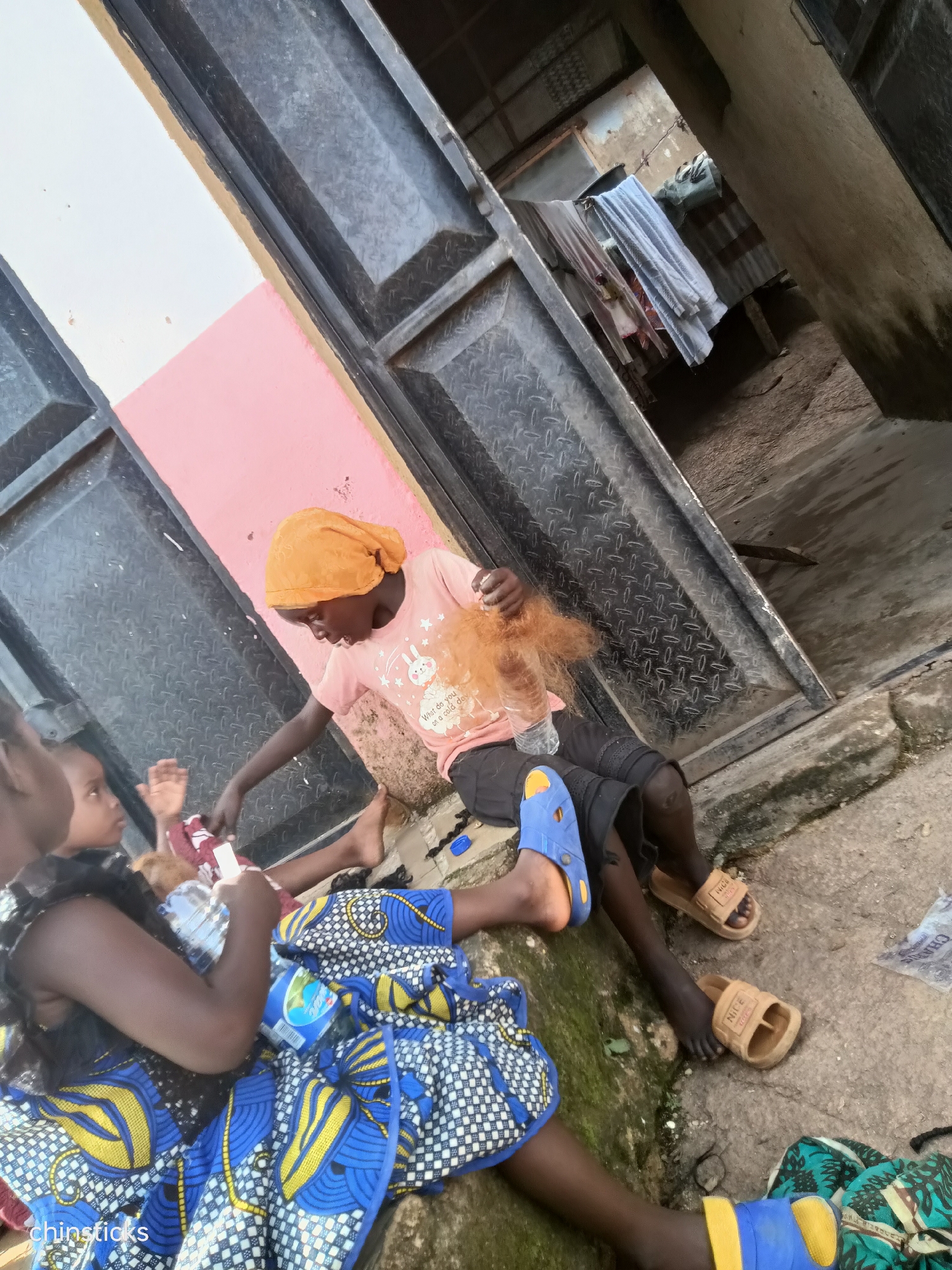 In Jos, Nigeria, an outdoor scene depicts several children in close proximity. The setting appears to be a rough, outdoor environment with concrete flooring and a pink-and-white painted wall behind the subjects. Two children are prominently seated on the ground, facing each other. One child, wearing a bright orange headscarf and a light pink t-shirt with a rabbit graphic, is holding a bundle of reddish-brown hair. This child is also wearing black shorts and blue and brown sandals. To the left of this child, another child, wearing a blue and yellow patterned dress, is also seated. A third child is partially visible on the far left, also in the blue and yellow dress, with their back to the camera. The children appear to be engaged in some form of activity, possibly related to hair styling or play. In the background, there are large metal doors and some laundry hanging on a line, suggesting a residential or semi-commercial area. The overall impression is one of everyday life in an urban Nigerian environment.