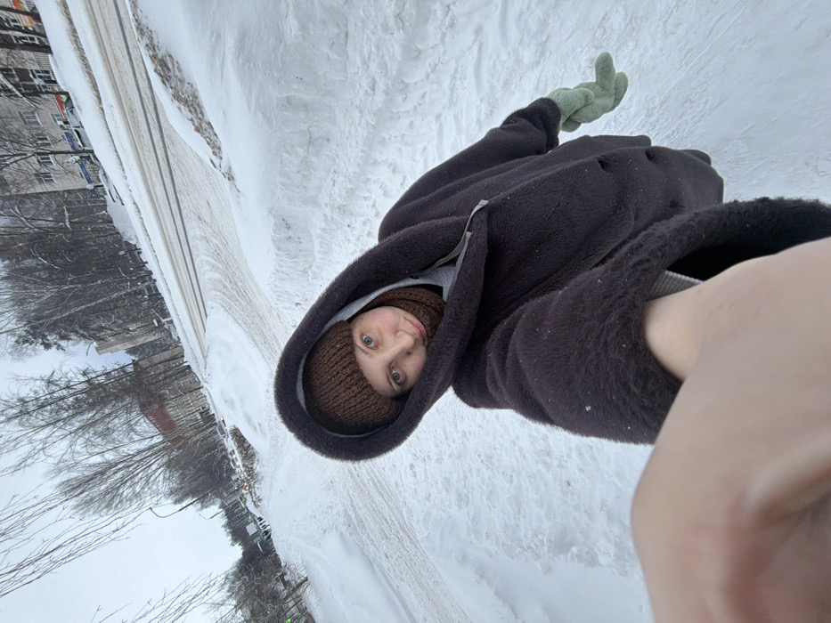 A female individual is depicted in a selfie, taken outdoors in a snow-covered urban environment within Chaikovsky, Russia. The subject occupies the foreground, facing the camera with a neutral expression, her left hand partially visible as she holds the camera. She wears a dark brown, textured hooded coat, a brown knitted beanie, and a matching brown knitted neck warmer. A light green glove is visible on her right hand. The background features a two-lane asphalt road, partially cleared of snow and exhibiting tire tracks, bordered by substantial snowdrifts on the sidewalks. Beyond the road, bare deciduous trees and coniferous trees are visible, intermingled with several multi-story residential or commercial buildings typical of an urban street. The sky is overcast, indicating daylight conditions. Utility poles are present along the roadside. The scene is consistent with a winter setting in a residential or mixed-use area of Chaikovsky.