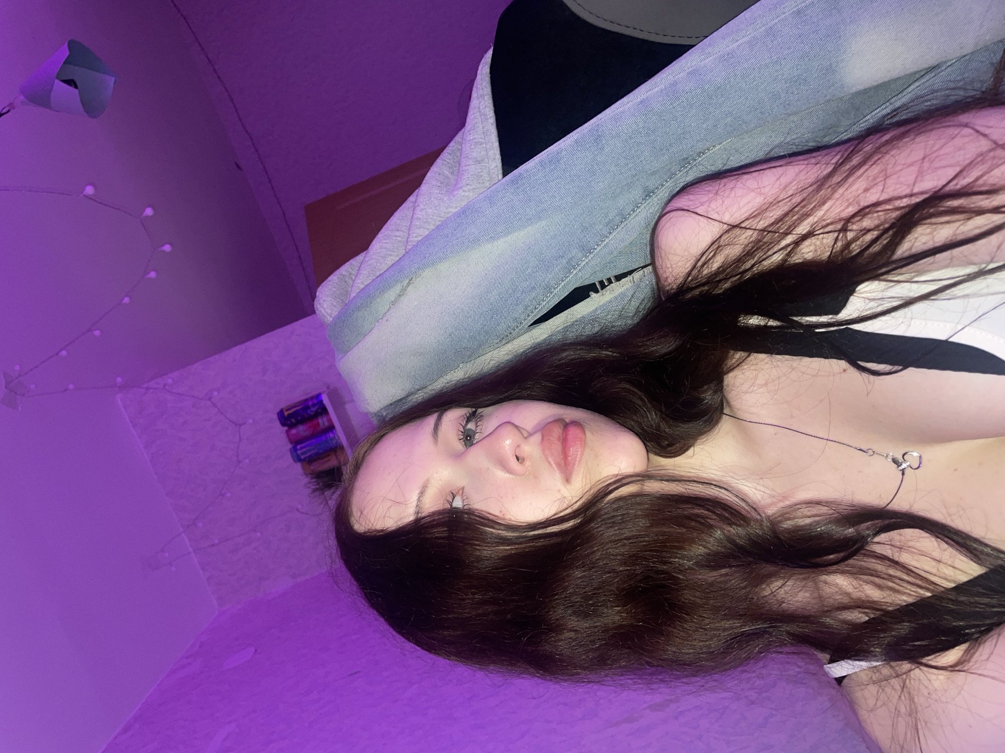 The image depicts a young woman with long, dark, wavy hair, light skin, and blue eyes, looking to her right. She is wearing a white top with black straps and a silver necklace with a heart-shaped pendant. She appears to be in a room with purple lighting, evidenced by the vibrant hue of the walls and ceiling. A string of small, round lights is visible across the ceiling. To her right, there is a crumpled object that appears to be made of denim fabric, possibly part of clothing or furniture. On a shelf in the background, several colorful cans, likely energy drinks, are arranged. The overall environment suggests an indoor, possibly residential, setting illuminated by colored lighting. The image does not contain any information that would allow for its location to be identified as Votkinsk, Russia.