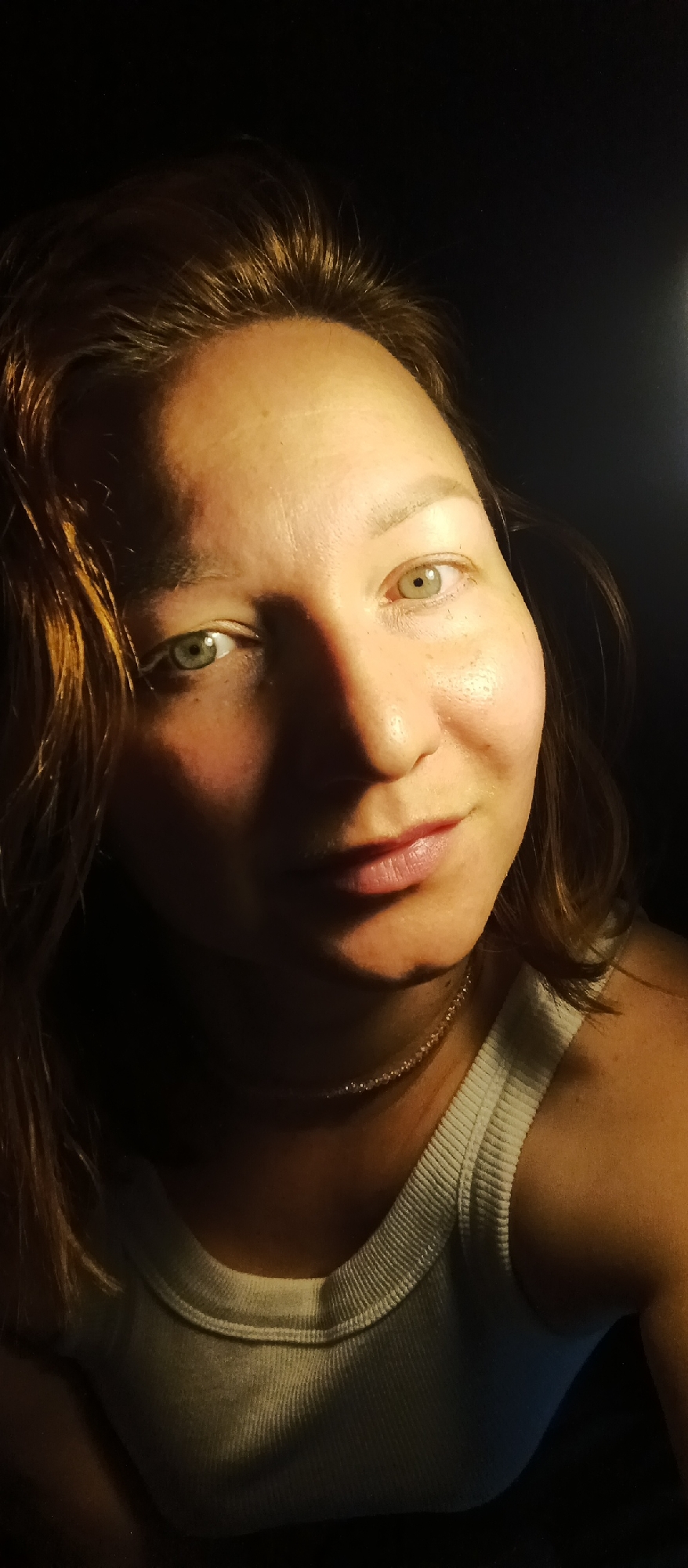 A close-up portrait depicts a woman with light skin and green eyes, taken against a dark background. The left side of her face is illuminated, highlighting freckles and skin texture, while the right side is cast in shadow. She has light brown hair and is wearing a white ribbed tank top and a delicate necklace. The location is Unknown, Unknown.