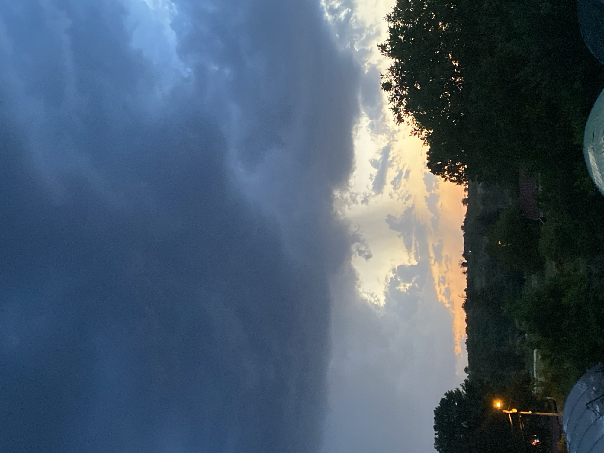The image shows a twilight scene in Konstantinovo, Bulgaria. Dark storm clouds dominate the left side of the frame. To the right, the sky transitions to a mix of orange and grey clouds, with visible sun rays breaking through. Silhouetted trees line the right edge of the image, with glimpses of buildings and vegetation interspersed. A street lamp glows in the lower right.