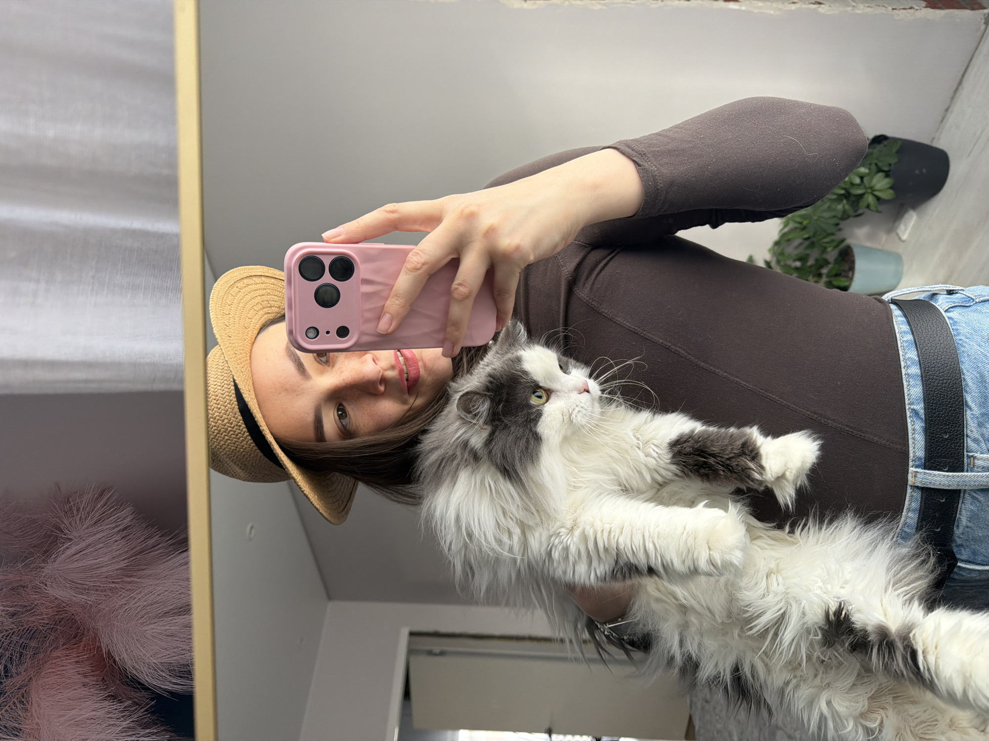 In a mirror selfie, a young woman is seen holding a fluffy, long-haired cat. The image is taken indoors, likely in a home in Chaikovsky, Russia, during the daytime, given the bright illumination.
The woman is partially visible, showing her head, right arm, and torso. She has dark hair and a pleasant, subtle smile, with light freckles on her nose and cheeks. She is wearing a straw hat with a dark band, a fitted brown long-sleeved top, and blue denim jeans with a black belt. Her right hand holds a pink smartphone with a distinct camera bump featuring multiple lenses, pointing it towards the mirror to capture the reflection.
The cat, primarily white with grey patches around its head, back, and paws, is cradled in the woman's left arm. Its fur appears thick and soft, and it has bright, alert eyes, looking upwards and to the right, away from the camera.
Reflected in the background are various elements of the room. To the far right, two potted houseplants are visible – one in a dark grey/black pot and another in a light blue pot. On the far left, a soft, pink, feathery decorative item is reflected. The mirror itself has a thin golden frame. There is no visible text in the image.