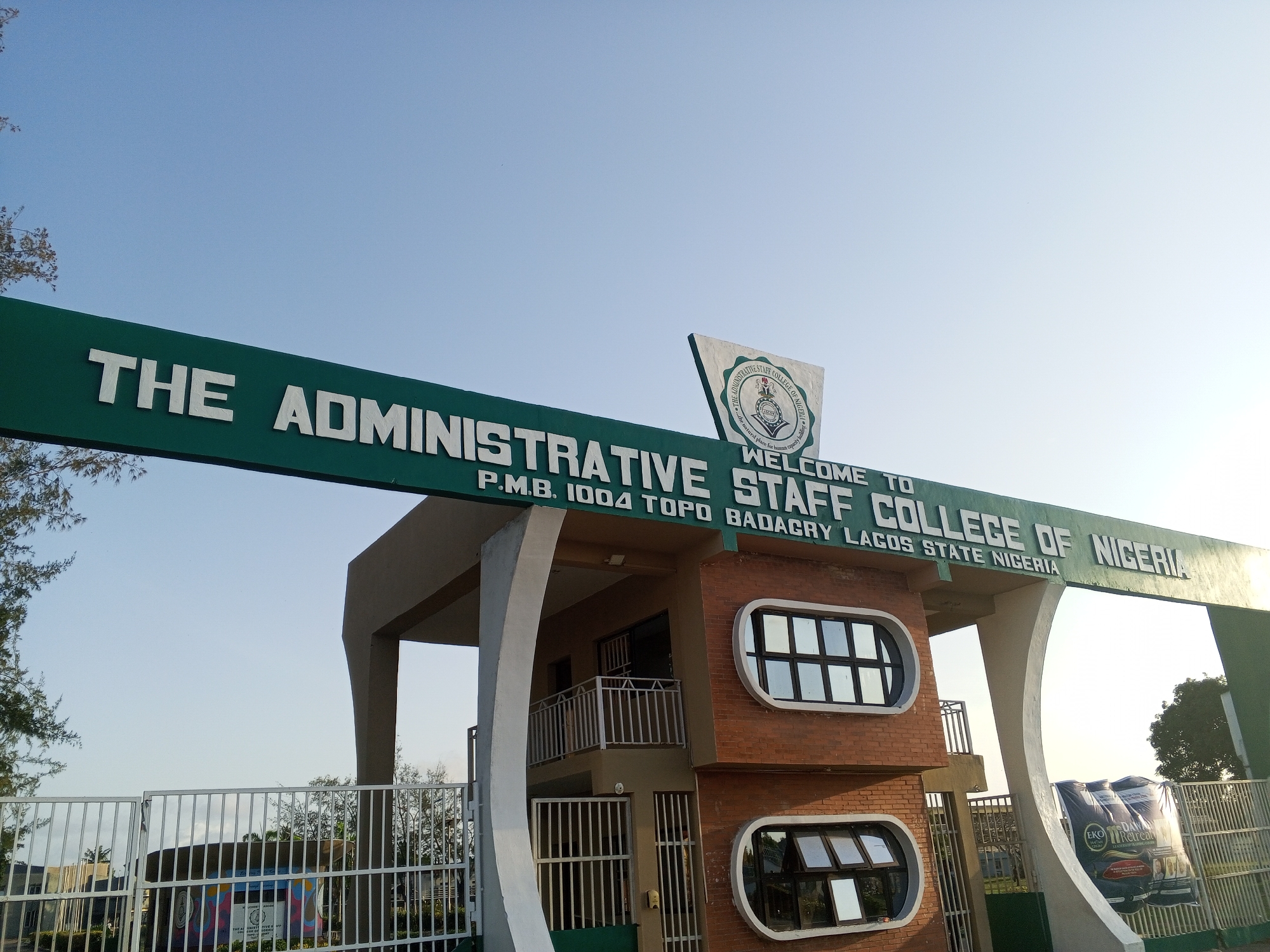 The image shows the entrance gate to The Administrative Staff College of Nigeria. The gate features a green overhead sign with white lettering. The address, "P.M.B. 1004 Topo Badagry Lagos State Nigeria" is also displayed on the sign. A seal is located atop the structure. The building is a brown brick structure with distinctive rounded windows. A metal gate is in the foreground. In the background, a banner that says "EKO Retreat" is visible. The location is in Topo, Nigeria.