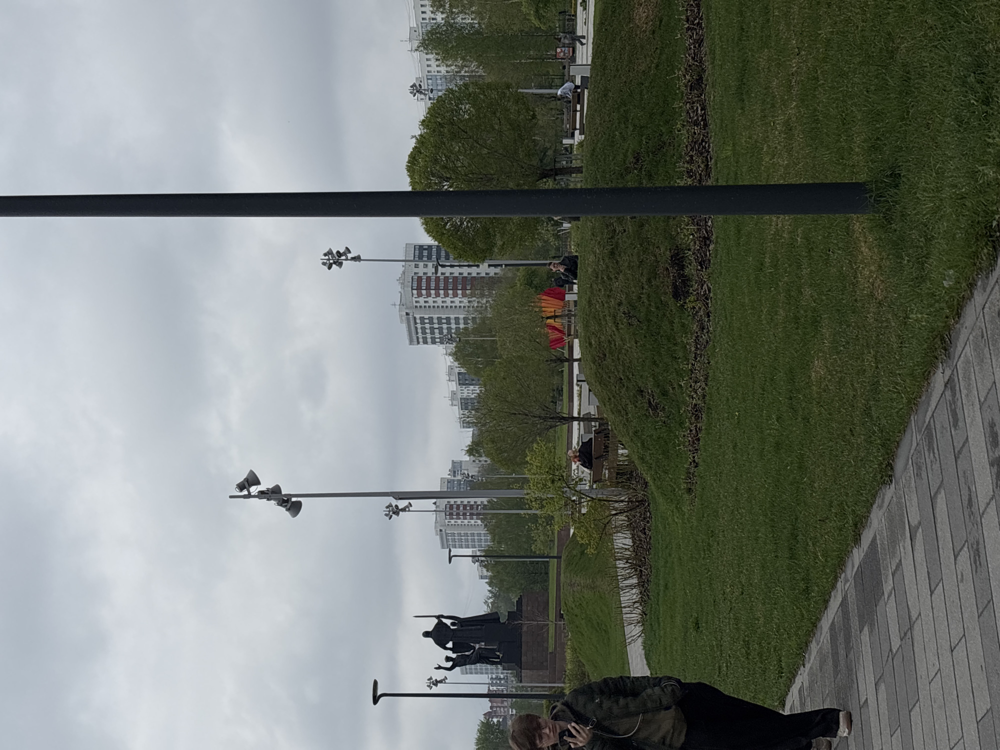 The image captures a park scene in Perm, Russia on an overcast day. A stone walkway runs along the right side of the frame, with a manicured green lawn to its left. A row of trimmed hedges separates the lawn from a line of benches and trees. Further back, residential buildings can be seen in the distance under a gray sky.
Several tall lampposts with multiple speakers are positioned along the walkway and within the park. A dark-colored monument depicting several figures stands along the walkway. In the foreground, a person is standing on the walkway and appears to be looking at their phone. A dark horizontal bar, possibly part of a structure or piece of equipment, stretches across the top portion of the image.