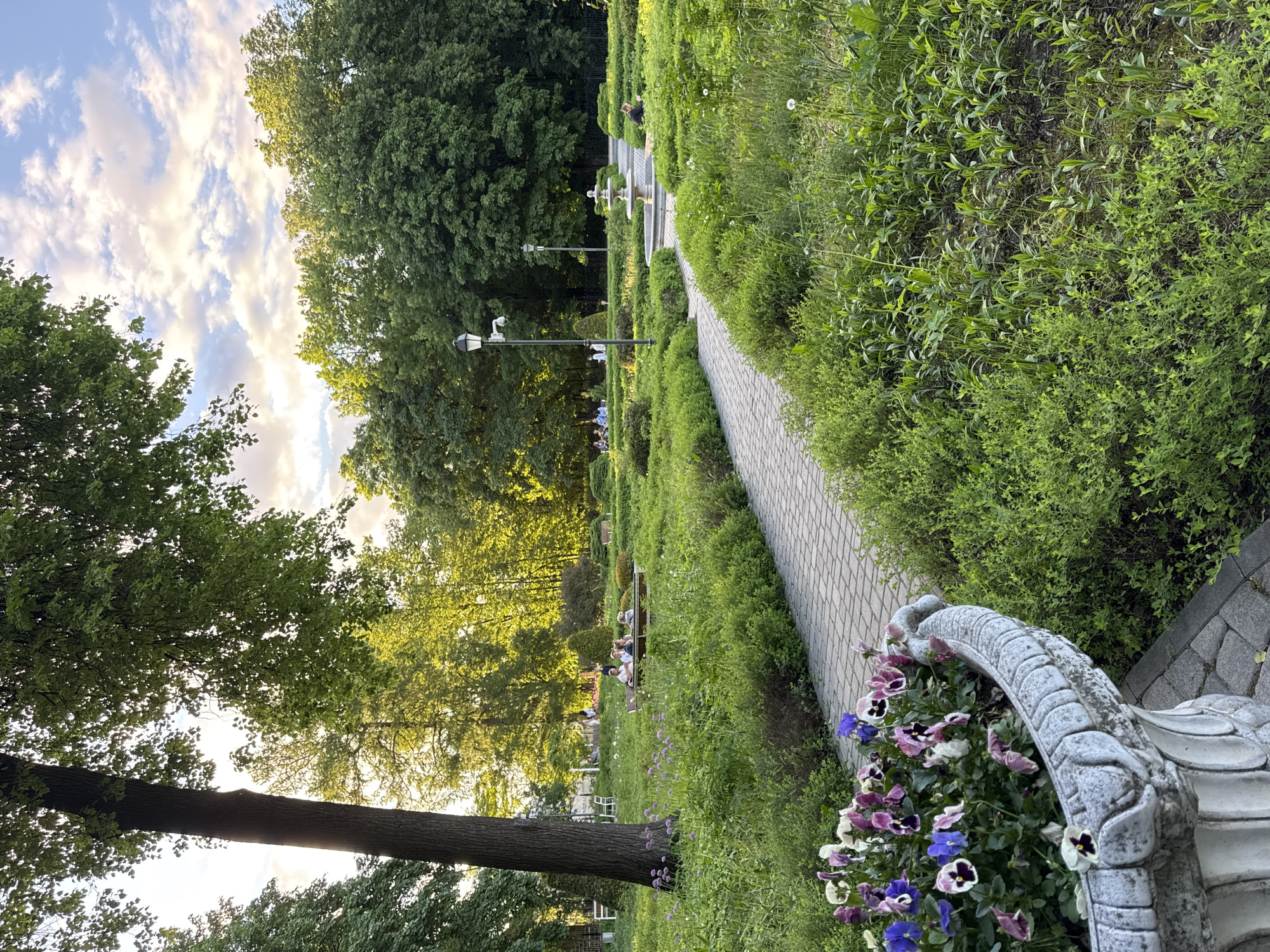 The image captures a serene scene in a park in Moscow, Russia. A stone-paved pathway meanders through manicured greenery, with neatly trimmed bushes lining both sides. Street lamps stand intermittently along the path. In the foreground, a decorative stone planter overflows with pansies in various shades of purple, blue, and white. In the background, people are seated on benches, enjoying the tranquil environment. Tall trees provide a backdrop, their green leaves creating a lush canopy. The sky is partially visible through the trees, revealing a mix of blue and clouds.