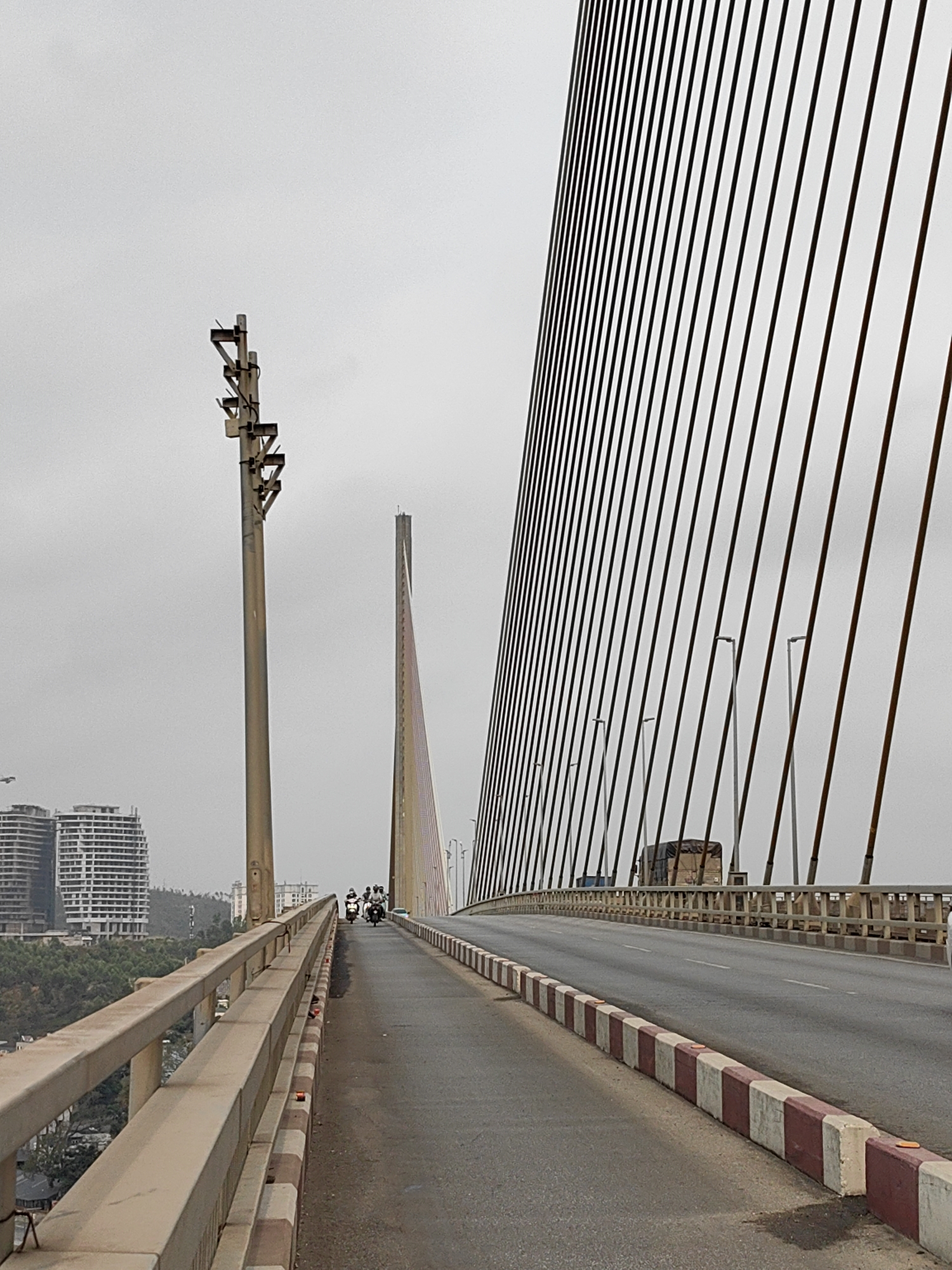 The image depicts a section of a cable-stayed bridge in Hong Gai Ward, Vietnam. The roadway is asphalt, flanked by concrete barriers with alternating red and white blocks. A utility pole is located on the left side. In the distance, motorcycles are moving along the bridge. Support cables anchor to the bridge deck, and a taller support tower is visible further down the span. A few high-rise apartment buildings can be seen on the horizon. The sky is overcast.