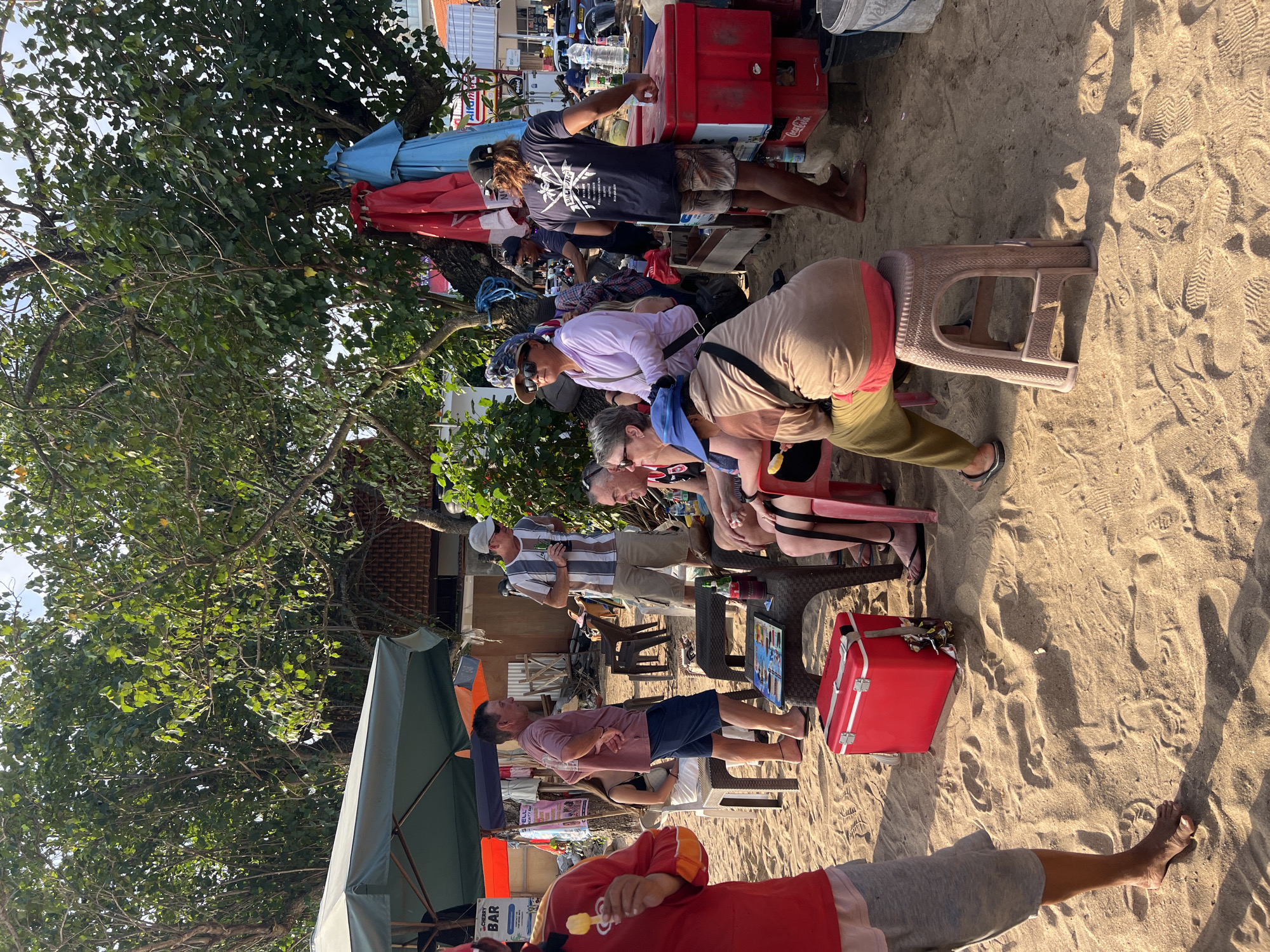 The image shows a group of people on a sandy beach in Kuta, Indonesia. The scene appears to be outdoors, likely a beachside bar or gathering spot, with several people seated and standing around, some holding drinks. Trees with dense green foliage provide shade. In the foreground, a person in a red jacket is holding an ice cream. Further back, several individuals are gathered around tables and coolers. Red and blue tarps are visible, along with umbrellas providing shade. The ground is covered in sand, with footprints indicating foot traffic. The overall atmosphere suggests a relaxed, casual gathering.