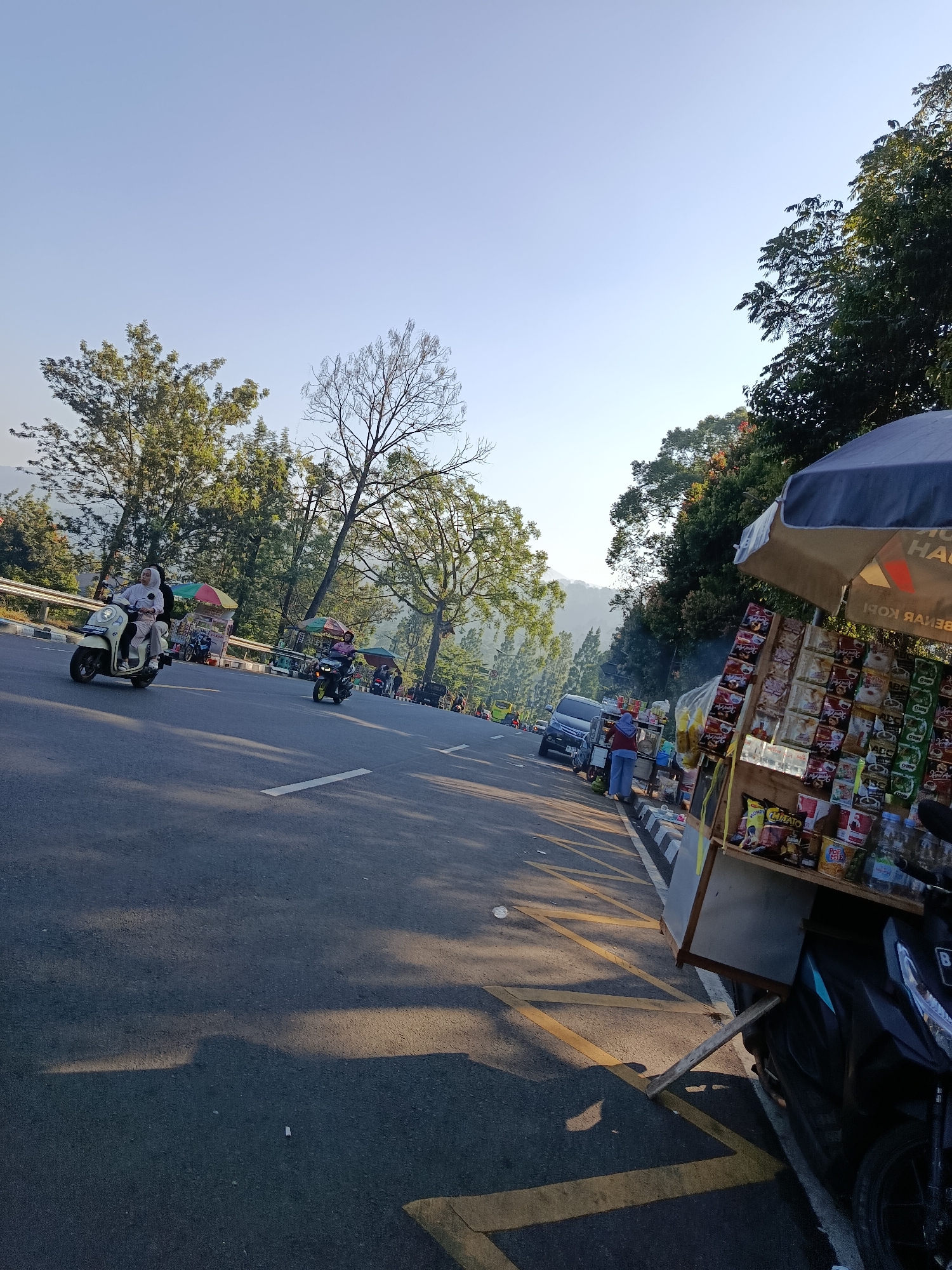 The image depicts a street scene in Cisarua, Indonesia, characterized by a paved road, lush green trees, and a clear blue sky. The road appears to be a two-lane thoroughfare with a white line marking the center and yellow lines at the edge. On the right side of the road, a small roadside stall is set up under a blue and grey umbrella, displaying various packaged snacks and drinks. A person, wearing a blue shirt and light-colored pants, stands behind the stall, appearing to attend to customers or manage the merchandise.
Several motorcycles are present on the road. One white scooter, with a rider wearing a light-colored hijab and trousers, is in the left lane, moving away from the camera. Another yellow and black motorcycle is also on the road, with a rider wearing a pink shirt. Further down the road, more motorcycles and a dark-colored car are visible, along with other indistinct figures and structures that suggest a bustling commercial area. The background is dominated by the natural landscape, with hills and dense vegetation framing the scene. The lighting indicates it is daytime, likely morning or late afternoon, given the long shadows cast on the road.