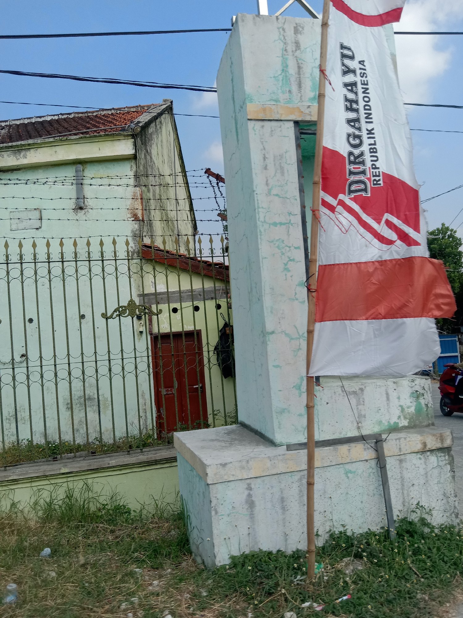 The image shows a partially completed monument or pillar with a flag of Indonesia attached. The flag is red and white, with the words "DIRGAHAYU REPUBLIK INDONESIA" written vertically in black. The monument appears to be made of concrete and is located outdoors, next to a light green building with a red door and a metal fence in front of it. The fence has barbed wire on top. The sky is blue with some clouds. The ground is covered in dry grass and some scattered debris. A red motorcycle is partially visible in the background. The scene is likely in Siwalan, Indonesia, based on the flag.