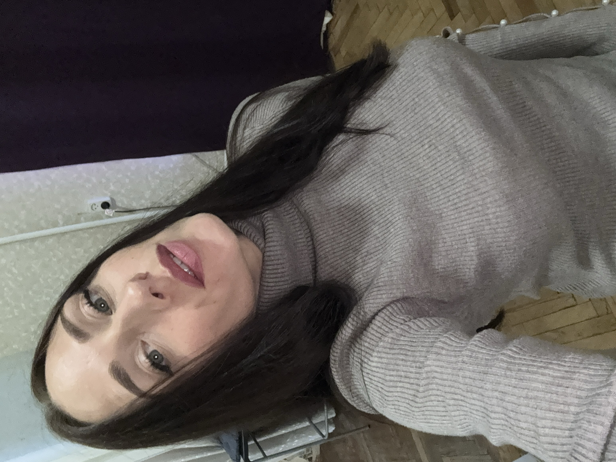 The image is a selfie taken indoors, likely in a residential setting in Saint Petersburg, Russia. A woman with dark hair and light skin is visible, looking directly at the camera. She is wearing a grey turtleneck sweater. The background shows a wall with a light-colored, textured wallpaper, an electrical outlet, and a dark curtain. The floor appears to be made of wooden planks. The lighting suggests an indoor environment, possibly during the daytime given the ambient light.