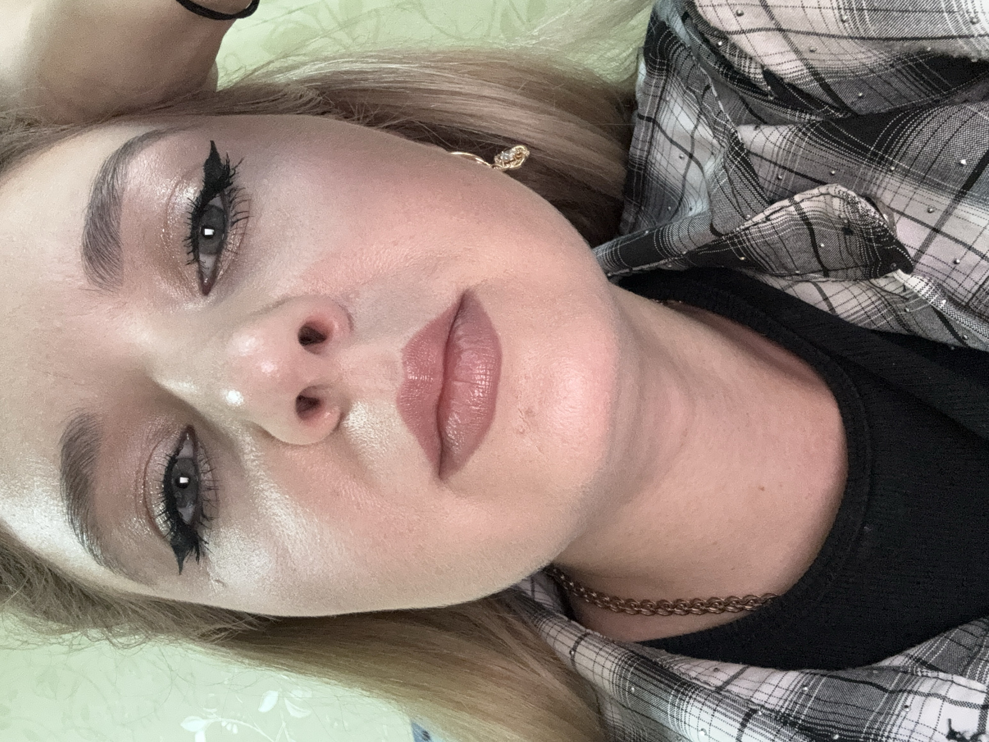 A close-up selfie shows the face of a woman with blonde hair, looking directly at the camera. She is wearing a black t-shirt and a plaid shirt with rhinestones. Her makeup includes winged eyeliner and shimmery eyeshadow. She has a gold chain necklace and a dangling earring on her right ear. The background is a light green wall with a faint floral pattern.