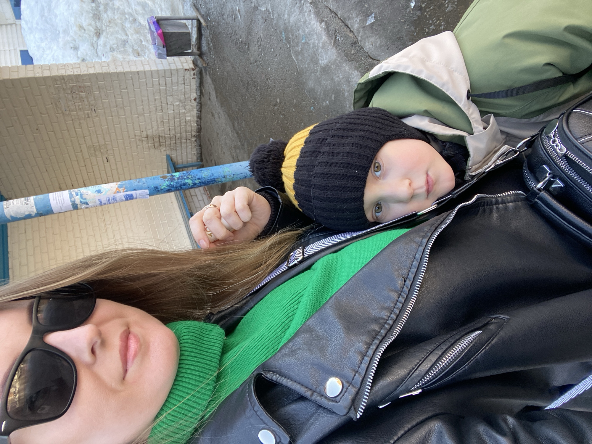 The image displays an adult female and a young child positioned at what appears to be a bus stop in the city of Noviy, Russia. The adult, visible from the chest up, wears black sunglasses, a green ribbed turtleneck, and a black faux leather jacket with silver hardware. A gold-toned ring is present on her right ring finger. The child, a male, is partially visible from the chest up, wearing a black and yellow knit beanie with a pom-pom and an olive green and beige hooded winter jacket; he is leaning against the adult's left side.
The immediate background features a weathered blue metal pole with peeling paint and several affixed flyers containing Cyrillic text. Behind the pole is a light beige brick wall, and a segment of a blue bench is visible. To the right of the wall stands a dark gray metal trash can containing a purple plastic liner. The ground is wet asphalt or concrete. Large, irregular piles of melting snow are visible behind the trash can and to the right of the structures, indicating a cold weather environment transitioning to warmer conditions.