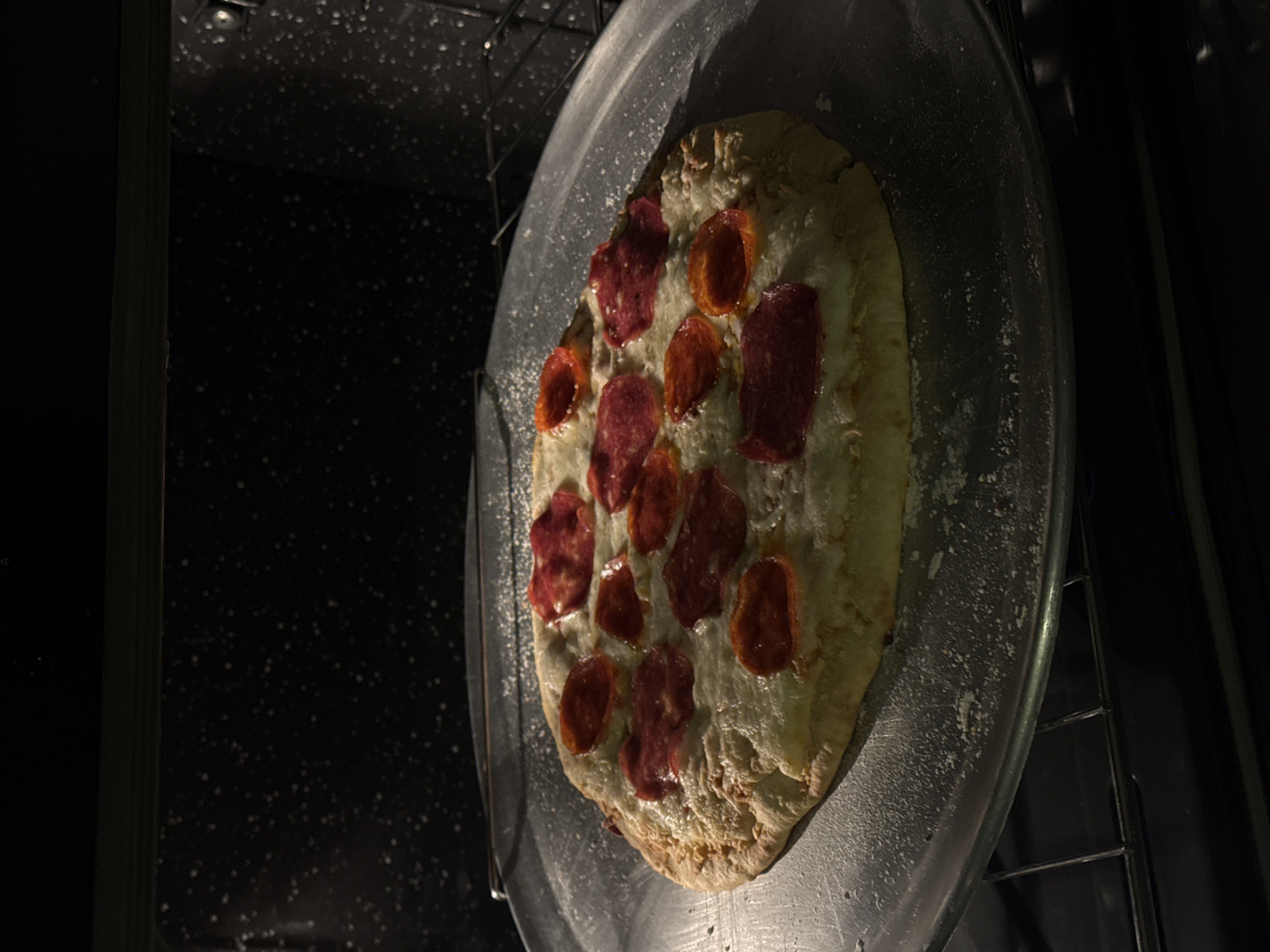 A pizza sits on a metal pizza pan inside an oven. The pizza has a light-colored crust, white cheese, and a mix of red and orange pepperoni slices. The oven interior is dark with speckled surfaces. The pizza pan rests on an oven rack. The image does not provide sufficient information to determine the location within San Bernardino Tlaxcalancingo, Mexico.
