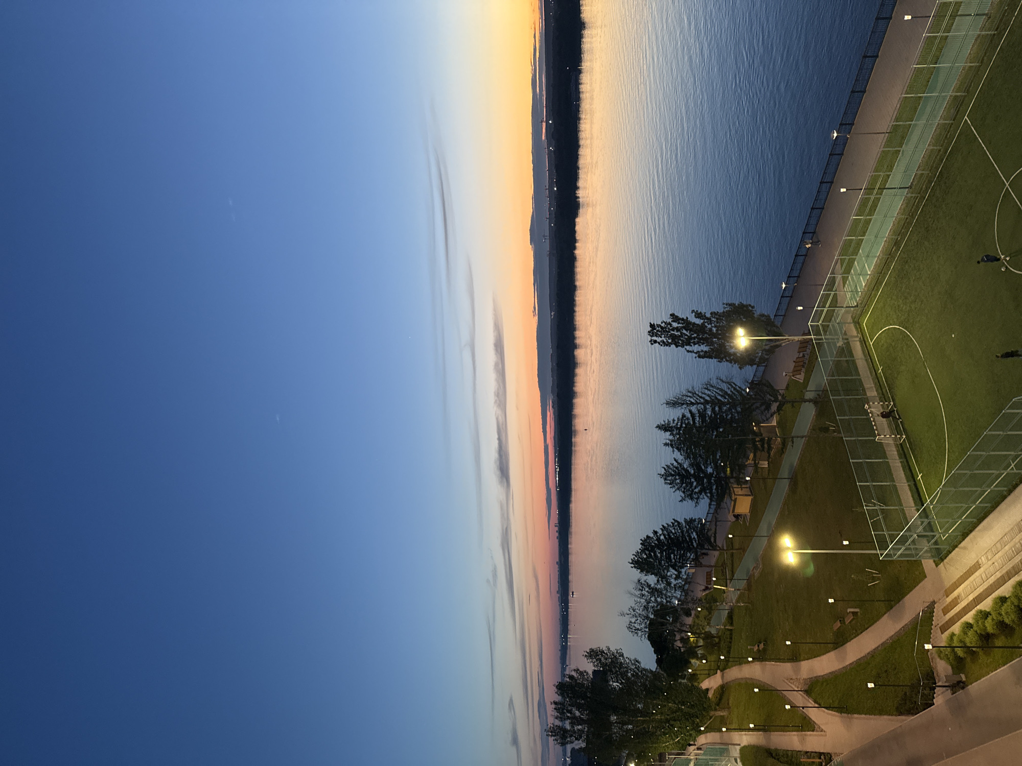 The image presents an elevated view of a public space at nightfall. To the left, a gradient of twilight sky transitions from deep blue to a pale orange sunset reflected in the water on the right side. A narrow landmass with indistinct features divides the sky from the water. Along the water's edge is a paved walkway bordered by a green space and a sports field. Lampposts line the walkway and illuminate the park area. The sports field has visible white markings and appears to be in use by at least two people in the upper right corner. The park has winding footpaths, mature trees, and decorative shrubbery. The location can be identified as Perm, Russia, based on the cityscape visible on the horizon in the sunset and the park's design.