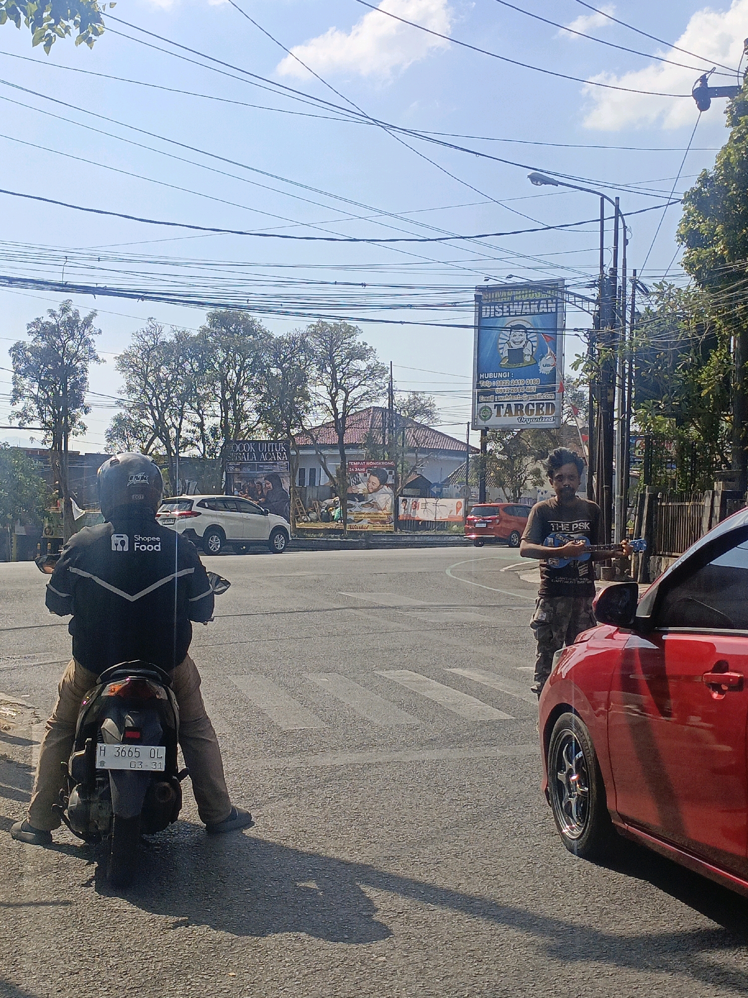 This image depicts a street scene in Salatiga, Indonesia, likely at an intersection due to the presence of a crosswalk and multiple vehicles. In the foreground, a person wearing a black jacket with "Shopee Food" branding is seated on a motorcycle, facing away from the camera. The motorcycle has a license plate displaying "H 3665 OL" and "03 - 31". To the right, a man stands on the side of the road playing a ukulele. He is wearing a t-shirt and camouflage pants. A red car is parked on the right side of the frame, partially obscuring the man. In the background, there are trees, buildings, and a large billboard. The billboard has text in Indonesian and features a cartoon illustration. Utility wires are strung across the sky, common in urban Indonesian settings. The lighting suggests it is daytime with clear skies.