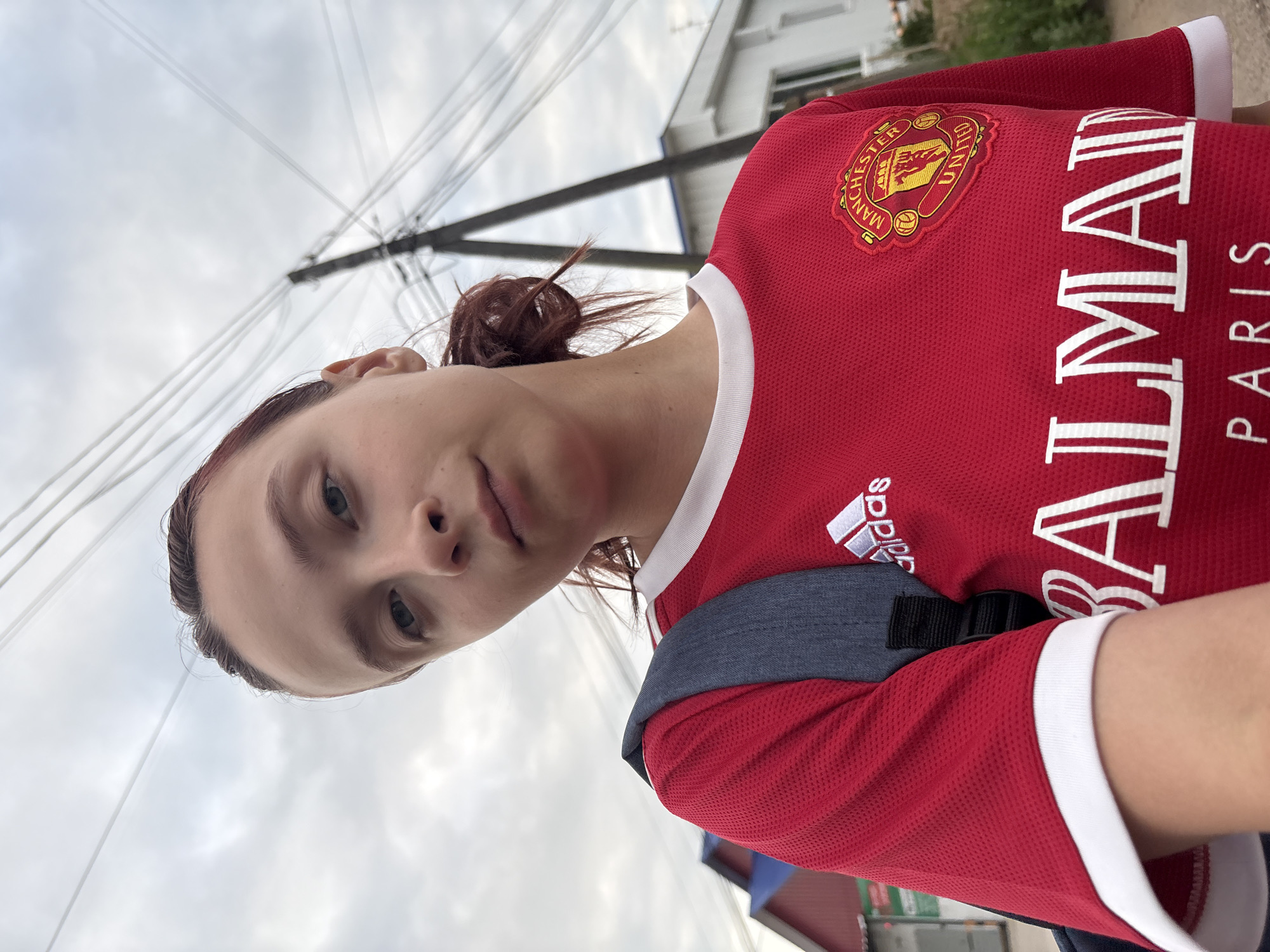 A young Caucasian female is pictured from the chest up, looking at the camera from a low angle. She has fair skin and her auburn hair is pulled back into a messy bun. She is wearing a red t-shirt with white trim around the collar and sleeves. The t-shirt has several logos, including an Adidas logo on the left chest and a crest with "MANCHESTER UNITED" on the right chest. The words "BALMAIN" and "PARIS" are printed in white letters across the front of the shirt. A grey backpack strap is visible over her left shoulder. The background is a slightly overcast sky with visible power lines and poles. To the left of the frame, part of a building with a red roof is visible. The location is identified as being in the city of Prikamskiy, Russia.