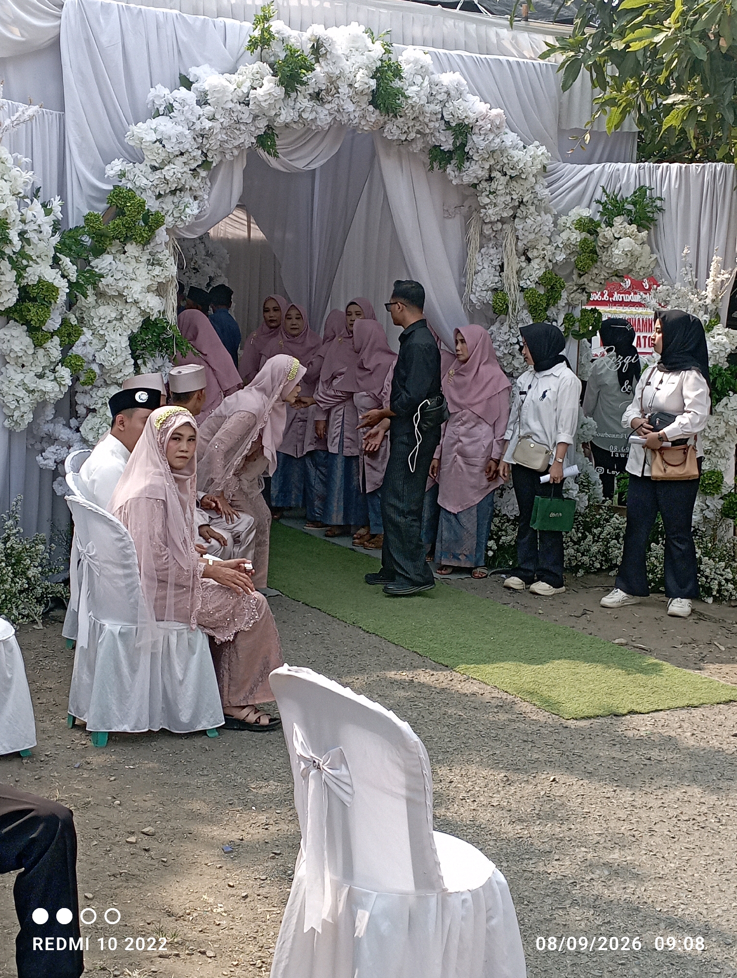 An outdoor wedding ceremony is taking place in Dukuhseti, Indonesia, with a floral archway and white drapes as a backdrop. A bride, adorned in a pink hijab and dress, sits next to a groom in a white shirt and cap. Several women in pink hijabs and traditional attire stand nearby, observing the ceremony. White chairs are arranged on the ground, and a green carpet runs down the aisle. The scene is illuminated by natural sunlight, casting shadows on the ground.