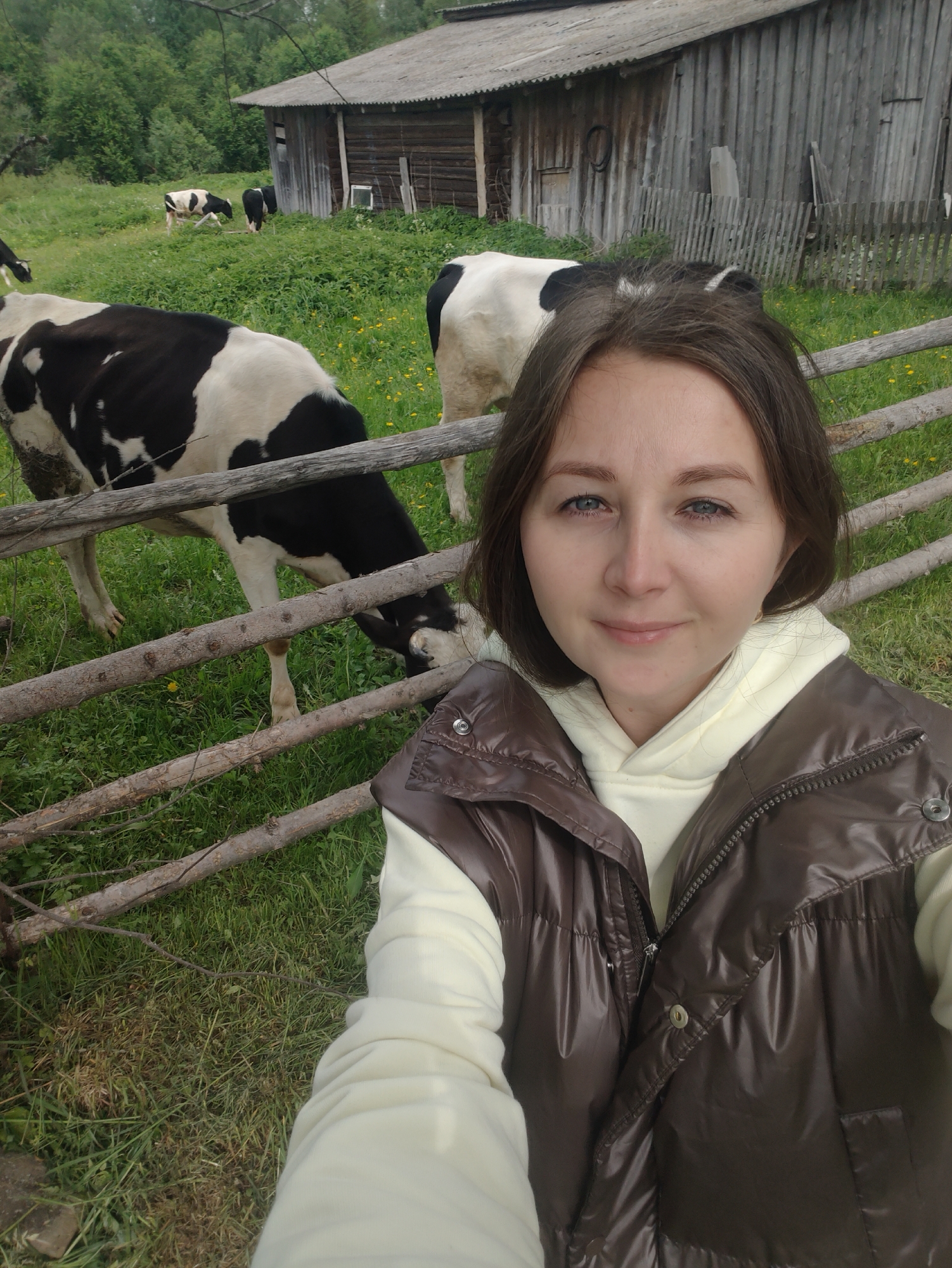 A woman in a cream-colored hooded sweatshirt and a brown vest is positioned in the foreground, slightly to the right. Behind her is a wooden fence, through which black and white cows are visible grazing in a green pasture. In the background, there is a wooden building with a weathered roof, surrounded by trees. The location appears to be a rural setting, potentially a farm or countryside.