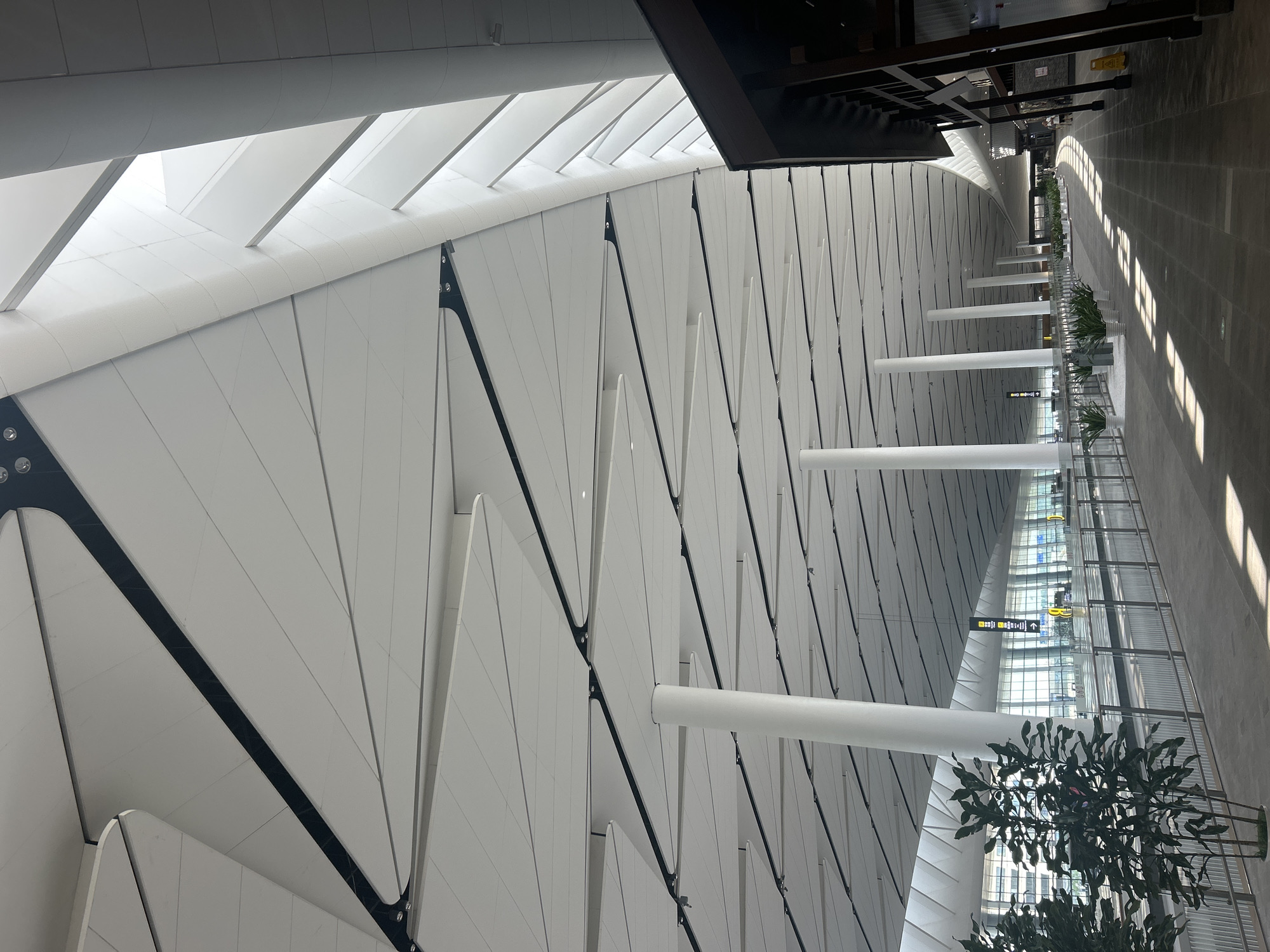 The image depicts the interior of a modern airport terminal, likely Jianyang City, China. The architectural style is characterized by a soaring, white, multi-paneled ceiling with dark accent lines, supported by numerous white cylindrical columns. Sunlight streams in from large windows along the far wall, creating dramatic light and shadow patterns on the tiled floor. A long, elevated walkway runs along the left side of the terminal, separated by a metal railing, and lined with potted plants. Signage, including directional arrows, is visible in the distance. The right side of the image shows a more traditional-looking building facade with dark wood elements and open doorways, suggesting retail or dining establishments. A few potted plants are also present on this side. The scene is devoid of any visible activity; there are no people or vehicles captured in the frame.
