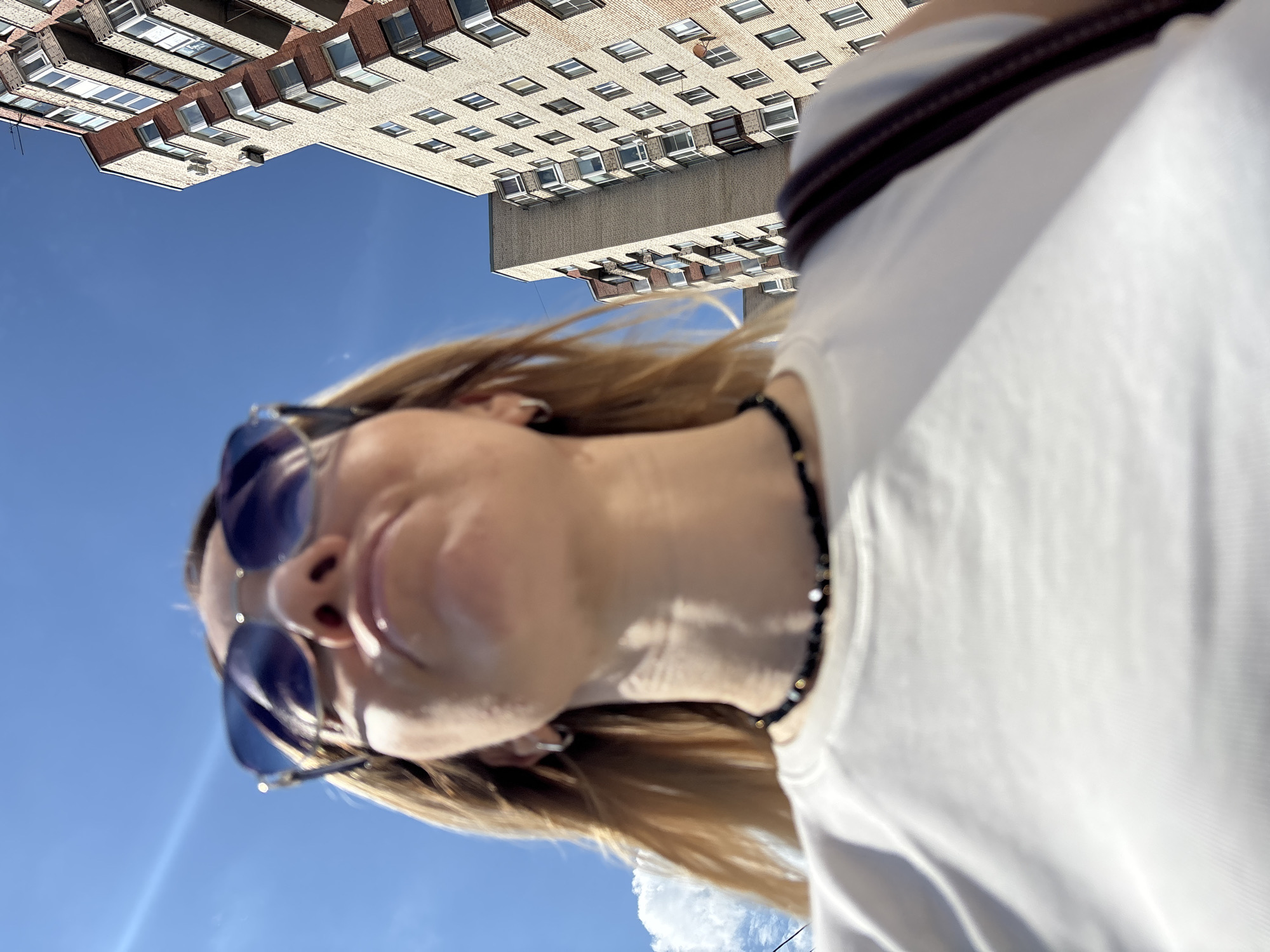 An outdoor, low-angle photograph captures a person looking upwards towards a tall building against a clear blue sky. In the foreground, the upper body and face of a person with blonde hair are visible, though partially out of focus. They are wearing blue-tinted aviator-style sunglasses, a black beaded necklace, and a plain white t-shirt. A dark, possibly brown or black, strap from a bag or garment is visible over their shoulder.
The background is dominated by a large, multi-story building constructed primarily of light beige or yellowish brick, with sections of darker brown brick or concrete detailing around some windows. The building features numerous rectangular windows arranged in orderly rows. The bright sunlight casts distinct shadows on the building's facade, indicating a clear, sunny day. The sky is a vibrant blue, with a small patch of white cloud visible towards the bottom right of the frame. The overall perspective suggests the person is standing close to the building and looking up, perhaps taking a selfie or being photographed from below. The architecture of the building is characteristic of urban environments, consistent with a city like Saint Petersburg, Russia. There is no visible text in the image.
