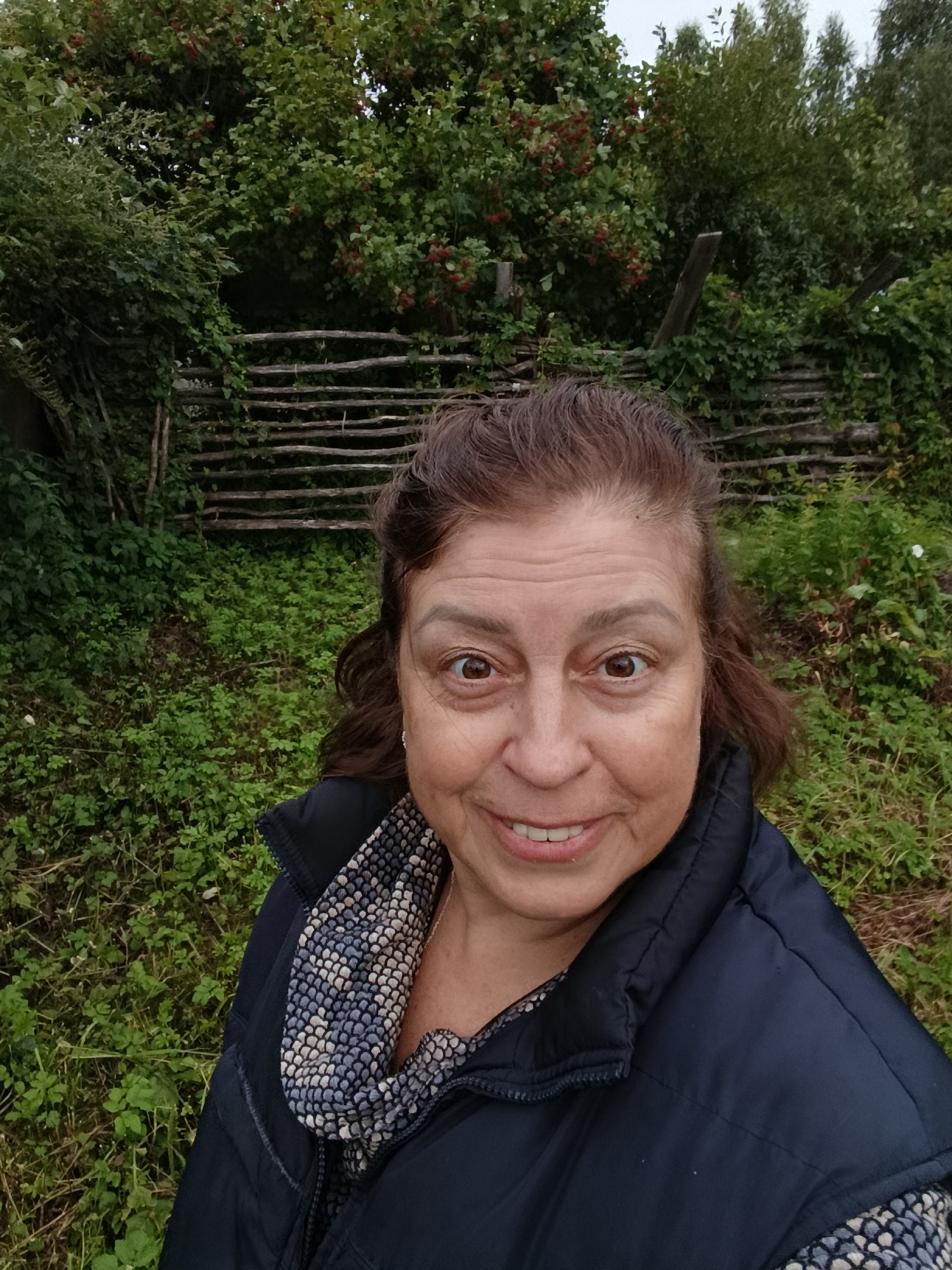 A woman with brown hair and brown eyes is standing in front of a wooden fence and lush green foliage. She is smiling and wearing a dark blue vest over a patterned scarf and a dark jacket. The environment suggests an outdoor setting, possibly a rural area or garden, consistent with the location of Верея, Russia, known for its historical sites and natural landscapes. The wooden fence is constructed from horizontal logs and appears weathered, blending with the surrounding greenery. The foliage consists of dense bushes and trees with some red berries visible on the branches. The overall impression is a candid portrait taken in a natural, rustic environment.