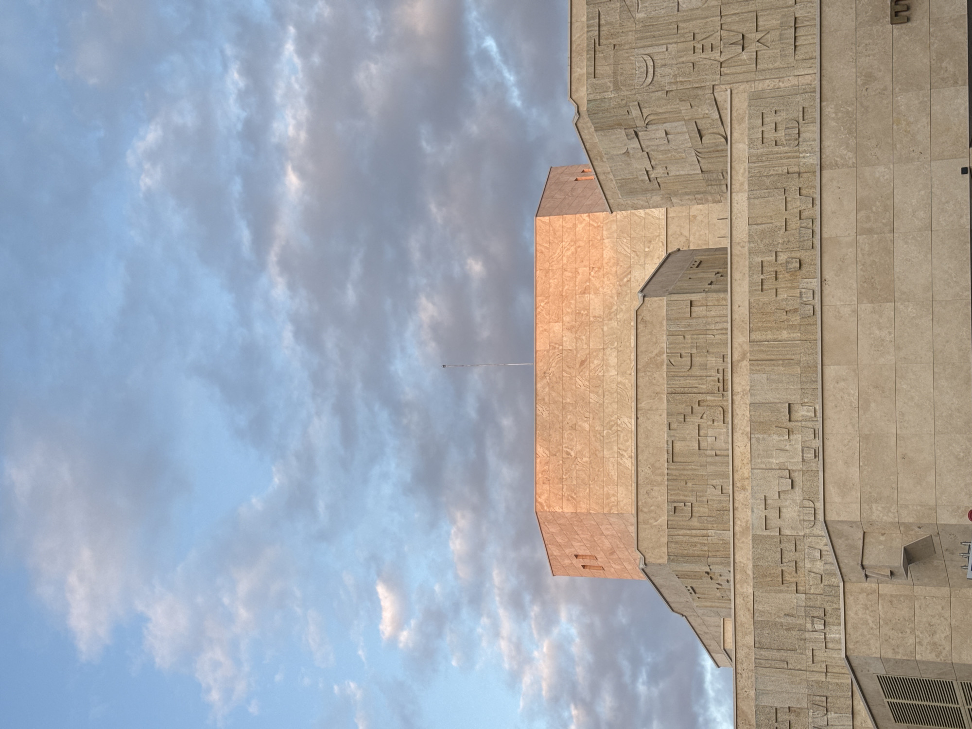 The image displays a monumental building facade against a partially cloudy sky, rotated 90 degrees clockwise, making the building appear on the right side of the frame and the sky on the left.
The primary subject is a large, multi-tiered building constructed from light-colored stone, likely travertine, exhibiting a distinctive architectural style. Its facade is heavily textured with elaborate, three-dimensional relief carvings and geometric patterns that resemble abstract script or symbols, creating a visually complex surface. The building is composed of various interlocking blocks, some of which catch a warm, golden-hour light, giving them a peachy-orange hue, while other sections remain in cooler beige and grey tones. A slender flagpole extends vertically from one of the central building sections into the sky.
The sky is a mix of soft blue and numerous white and grey clouds, some wispy and diffused, others denser, suggesting a partly cloudy day. The lighting on the building, with its warm highlights, indicates either early morning or late afternoon. No specific activity or event is taking place; the scene is a static architectural study.
Notable details include the intricate patterns covering the building's exterior, which are a prominent feature of its design. While these patterns might evoke lettering or hieroglyphs, no clearly legible text in any language is discernible on the main facade. On a far-right section, a small red circular object, possibly an alarm, is visible, with faint, unreadable text below it. Given the provided context, the building is located in Sofia, Bulgaria, and its distinctive design strongly suggests it is the National Palace of Culture (NDK).