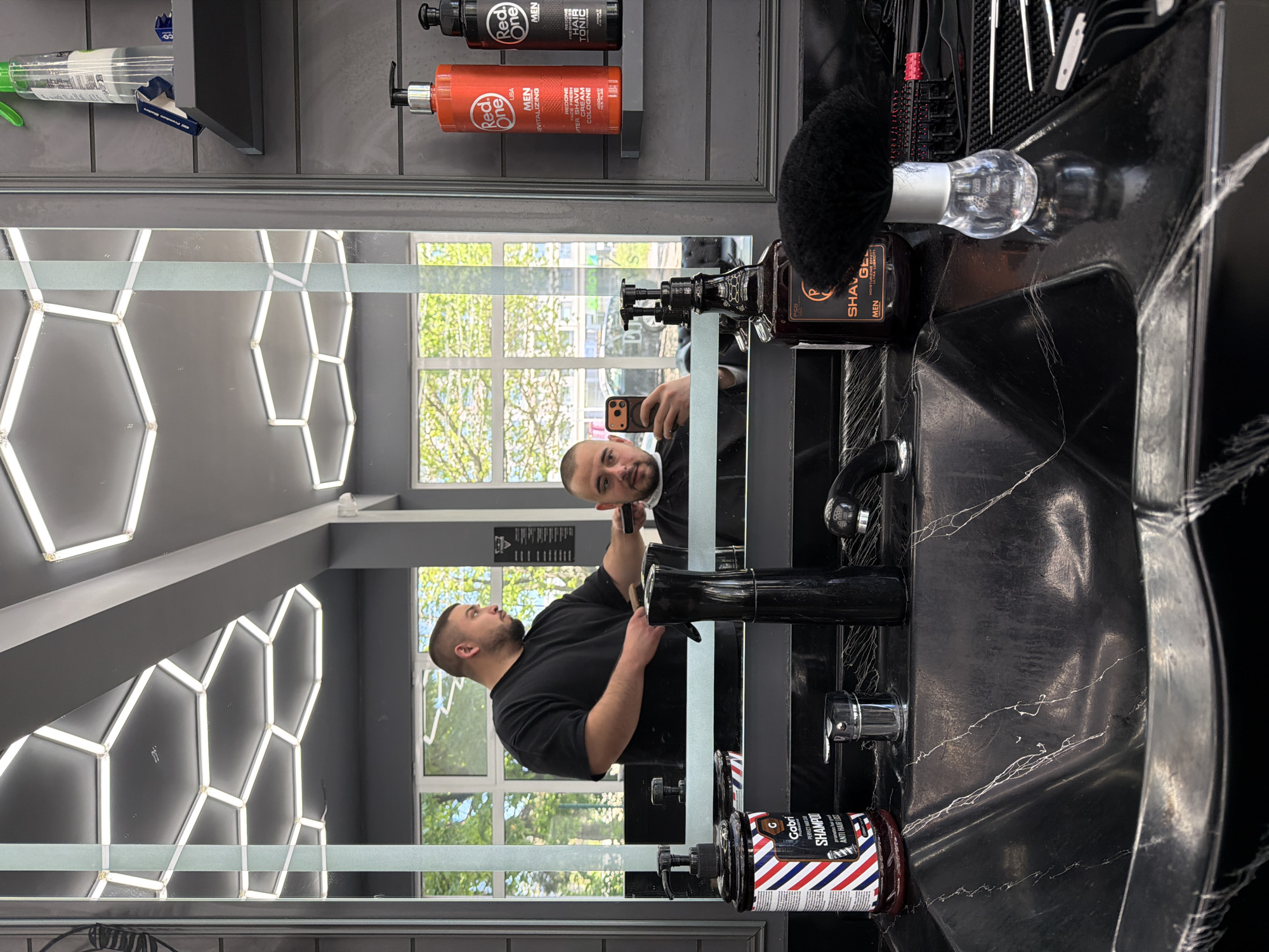 Here is a forensic description of the image:
**Scene:** A barber shop or salon. The image is taken in front of a large mirror.
**People:** A man in a black t-shirt is visible in the mirror. He is holding a mobile phone with a brown case and is taking a selfie in the mirror. He has a shaved head and a beard.
**Objects:** Several bottles of hair products are on shelves behind the mirror, including a red-labeled "Red One" cologne. A black bottle of shaving gel and a shaving brush with a transparent handle are placed on the black sink. A bottle of hair shampoo, branded "Gabri," can be seen on the left edge of the sink. The sink has a black marble-like finish with white veins. The mirror has a lighting fixture of hexagonal shapes behind it.
**Actions:** The man is taking a selfie in the mirror.
**Interactions:** The man is interacting with his reflection, taking a photo.
**Environment:** The room appears well-lit with natural light coming through windows and artificial light from the hexagonal light fixtures. The walls are painted in a gray color. The window has a view of the exterior, with a glimpse of trees and buildings, though the exact city or location cannot be determined. The general condition of the room appears neat and clean.