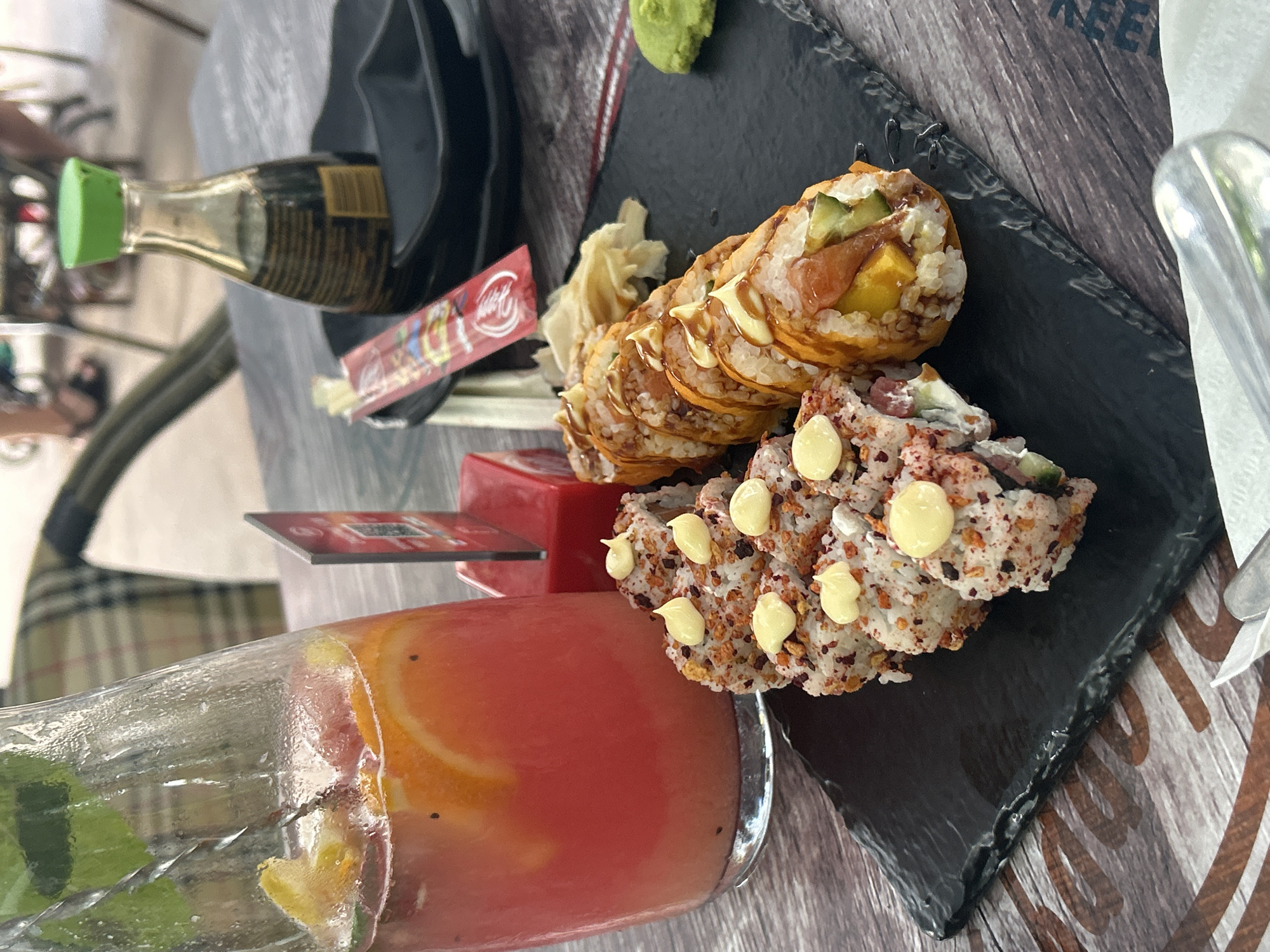 The image shows a table setting, likely in a restaurant in Burgas, Bulgaria. The scene includes a plate of sushi with multiple types of rolls, presented on a dark, possibly slate, platter. A glass of reddish-orange beverage, containing slices of orange and what appears to be mint, sits beside the sushi. Other visible items include a small bottle of soy sauce with a green cap, packaged chopsticks, a small container of wasabi, and a container of ginger. The table has a weathered, wood-like surface. Some blurry figures are visible in the background, suggesting the setting is an outdoor dining area or patio. The setting is likely informal and represents a meal in progress or prepared for consumption.