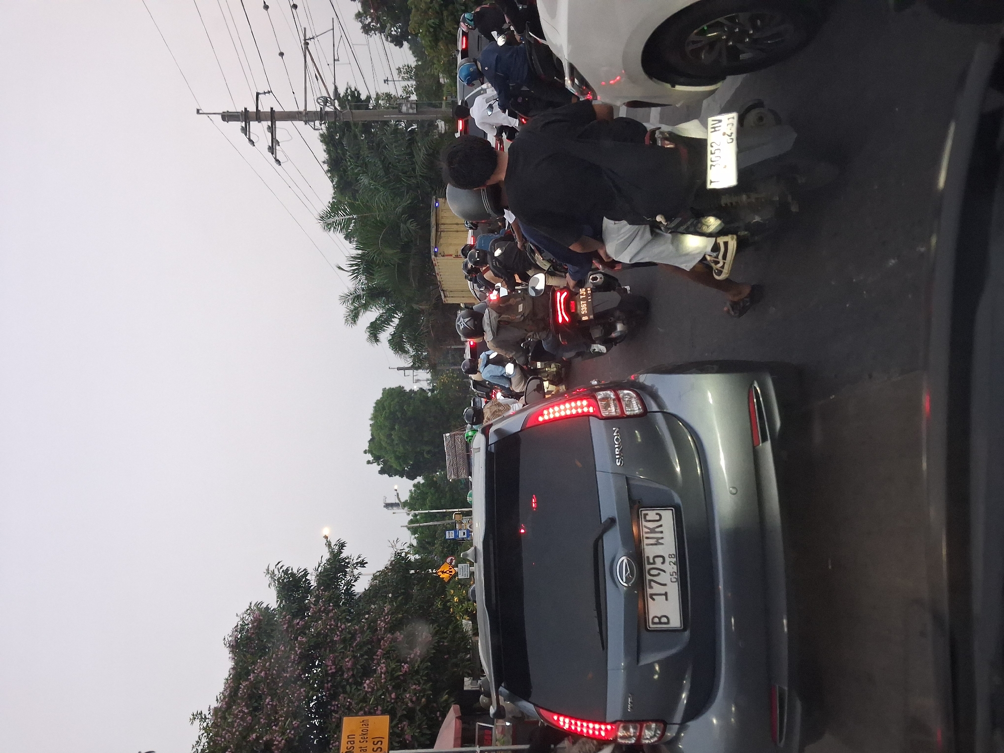 The image depicts a traffic jam on a street in South Jakarta, Indonesia. The sky is overcast, suggesting it might be late afternoon or early morning. The foreground is dominated by the rear of a gray hatchback car with a license plate reading "B 1795 WKC". Behind this car, a dense stream of motorcycles and a few cars are visible, all moving slowly or stopped. Many of the motorcycles have riders wearing helmets, and some are carrying passengers. The general environment appears to be urban, with trees and utility poles lining the street. A yellow sign with Indonesian text is visible on the left side of the image, partially obscured by trees. The scene conveys a sense of congestion and the typical daily commute in a busy city.