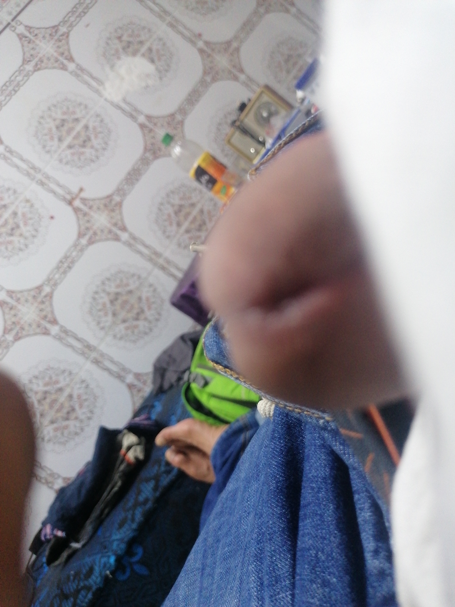 The image shows a close-up view of a person's hand and arm, in a relaxed pose, against a tiled floor. The floor has a repeating pattern of white tiles with a decorative, floral-like motif in shades of pink and brown. To the left of the hand, there is a black cloth bag with a blue floral pattern. A bright green backpack is partially visible behind the bag, with a white cord detail at the top. Further in the background, there is a bottle with a yellow label and a green cap, and what appears to be a framed picture or object. The overall impression is a casual, indoor setting, possibly a home or a common area, within the city of Essaouira, Morocco.