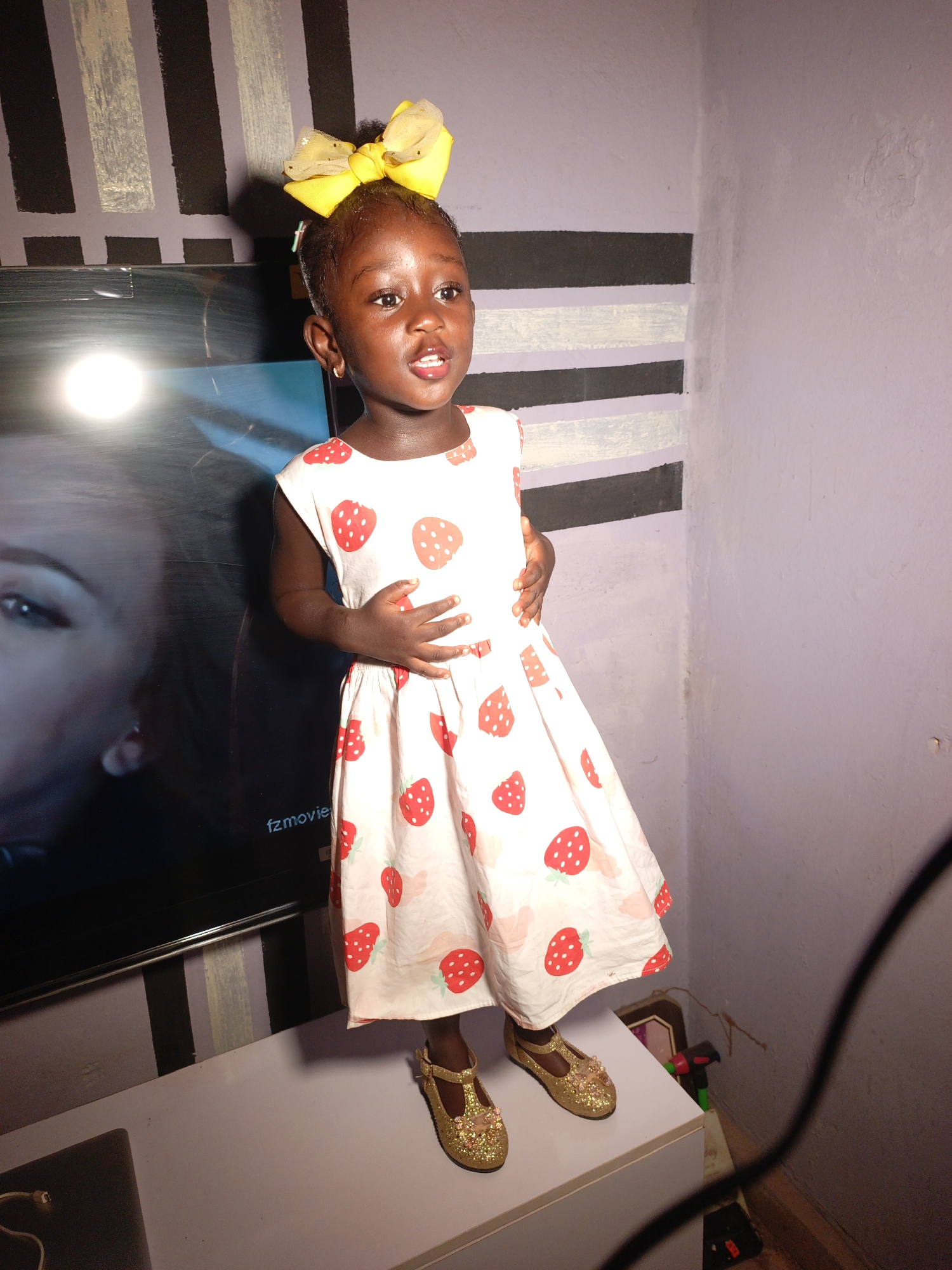 An indoor portrait features a young Black child, likely a girl, standing on a white dresser or cabinet. She is positioned slightly right of center, facing slightly to her right with her mouth slightly open, as if speaking or reacting. The child has dark skin, dark eyes, and short hair styled with a large, bright yellow bow. She wears a sleeveless white dress patterned with red strawberries and sparkly gold t-strap shoes.
To her left, a large flat-screen television is visible, displaying part of a human face (an eye, eyebrow, and nose) and the text "fzmovie" at the bottom center. A prominent bright, circular light reflection is on the upper left side of the TV screen.
The background shows the corner of a room. The left wall features horizontal stripes of black and light grey/white against a light purple base. The right wall is a solid light purple, showing some minor texture or wear. A black cable runs vertically along the right edge of the frame. In the bottom right corner, behind the dresser, indistinct objects like a green and black item and a framed picture are partially visible. The child's expression appears curious and engaged in this casual indoor setting in Akwanga, Nigeria.