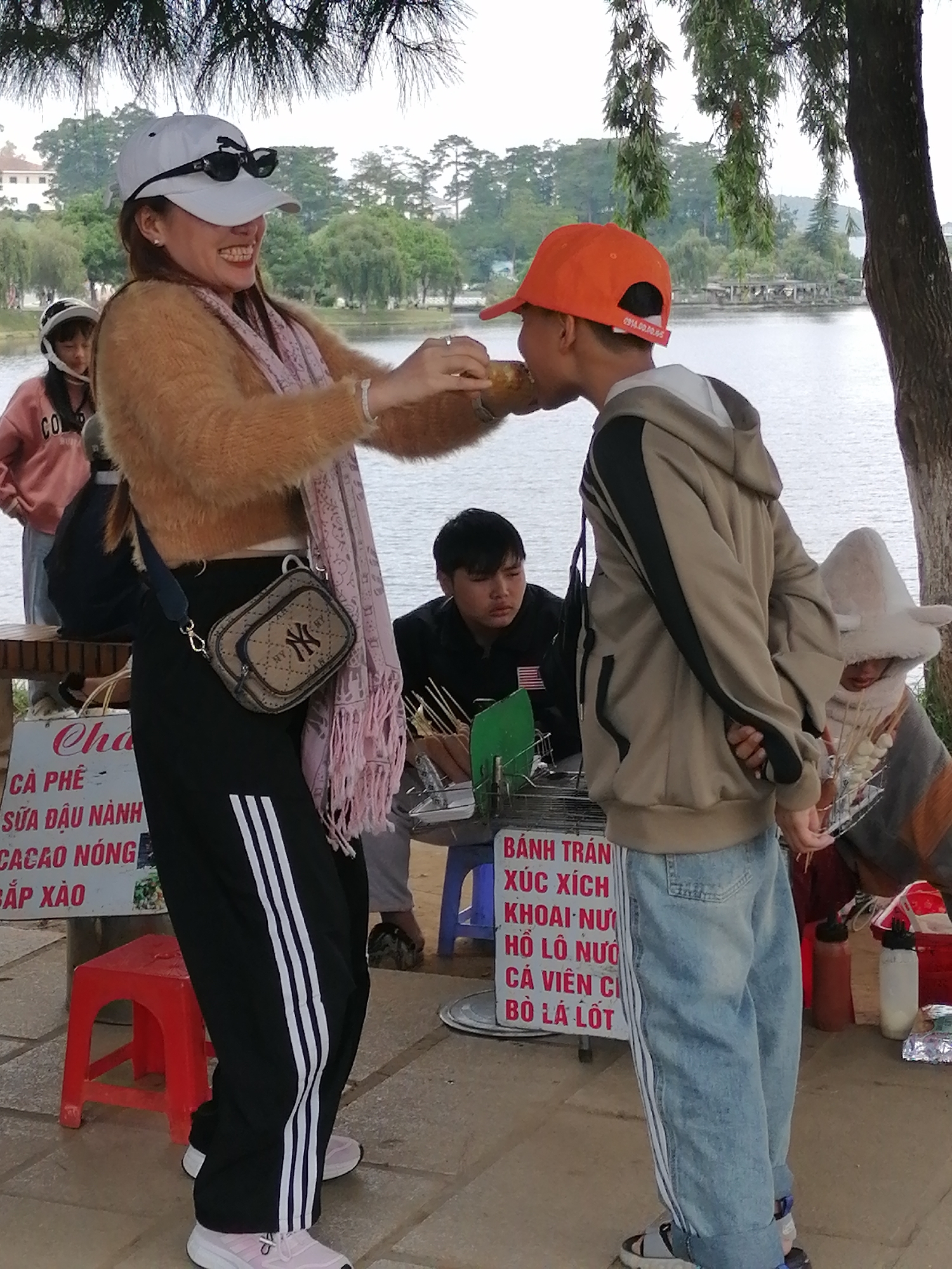 A woman wearing a white baseball cap, sunglasses, a fuzzy tan sweater, and black pants with white stripes is feeding a young boy wearing an orange baseball cap and a tan hooded jacket. The boy is holding a piece of food to his mouth. The woman is smiling and holding the food. In the background, there is a body of water, trees, and buildings. There is also a sign with Vietnamese writing that says "BÁNH TRÁM" which means "rice paper rolls." The scene appears to be at a street food stall near a lake in Da Lat, Vietnam. Other people are visible in the background, including a man sitting at a table and a person wearing a hooded hat. The ground is paved.