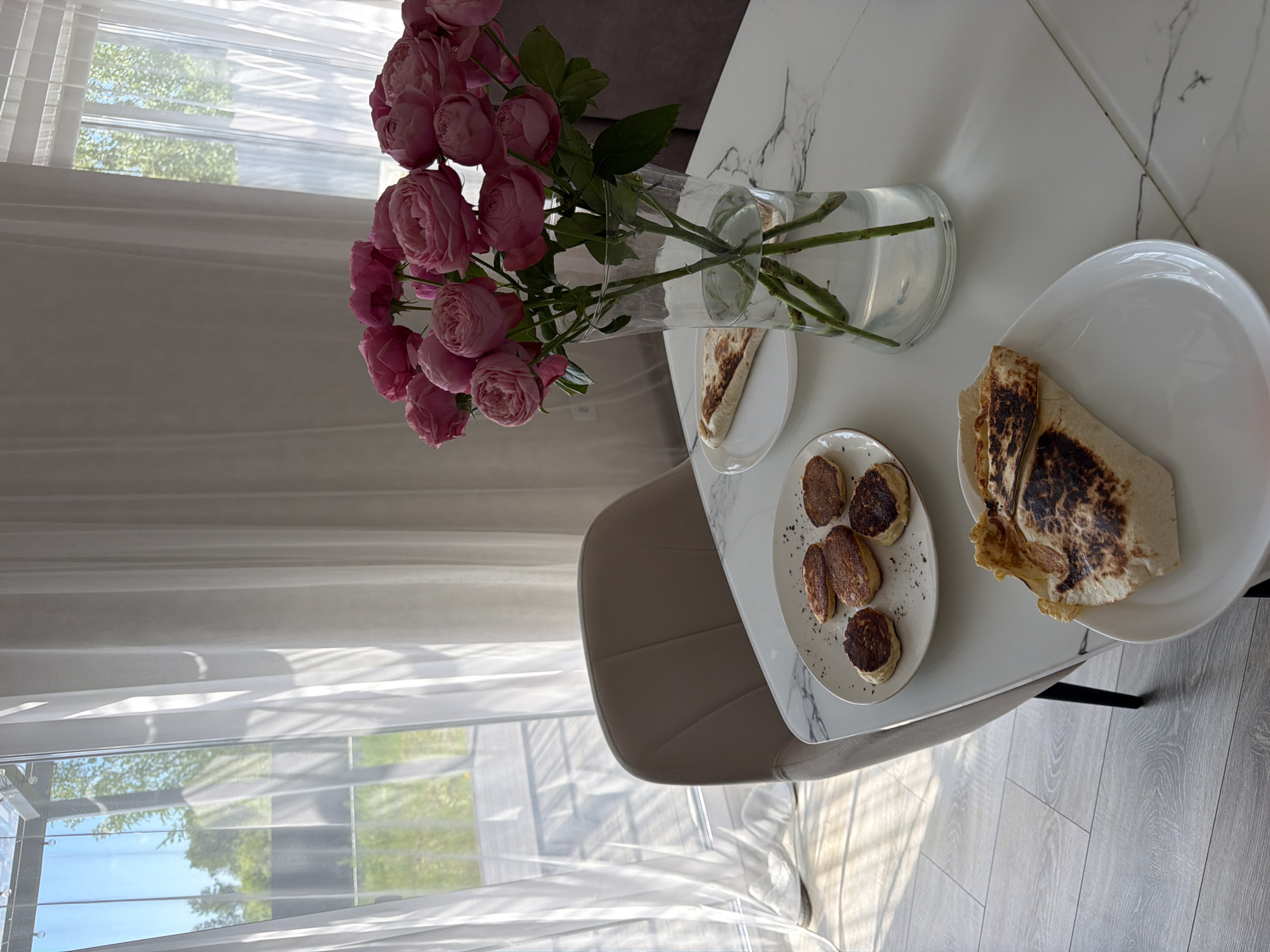 A still life image taken indoors, likely in Malinovka, Russia, features a dining table set for a meal. The table is white with a marble-like pattern and is placed near a window with sheer white curtains. Sunlight streams through the window, casting striped shadows on the floor.
On the table, a bouquet of pink roses in a clear glass vase is prominently displayed. To the left of the vase, a white plate holds several small, round pancakes, some appearing slightly burnt. Another white plate in the foreground displays a folded flatbread with grill marks. A single folded flatbread is also visible on a smaller white plate behind the vase.
A beige upholstered chair is positioned at the table. The flooring appears to be light-colored wood planks. The background suggests an outdoor environment visible through the window, with greenery and a fence. No people are present in the image.
