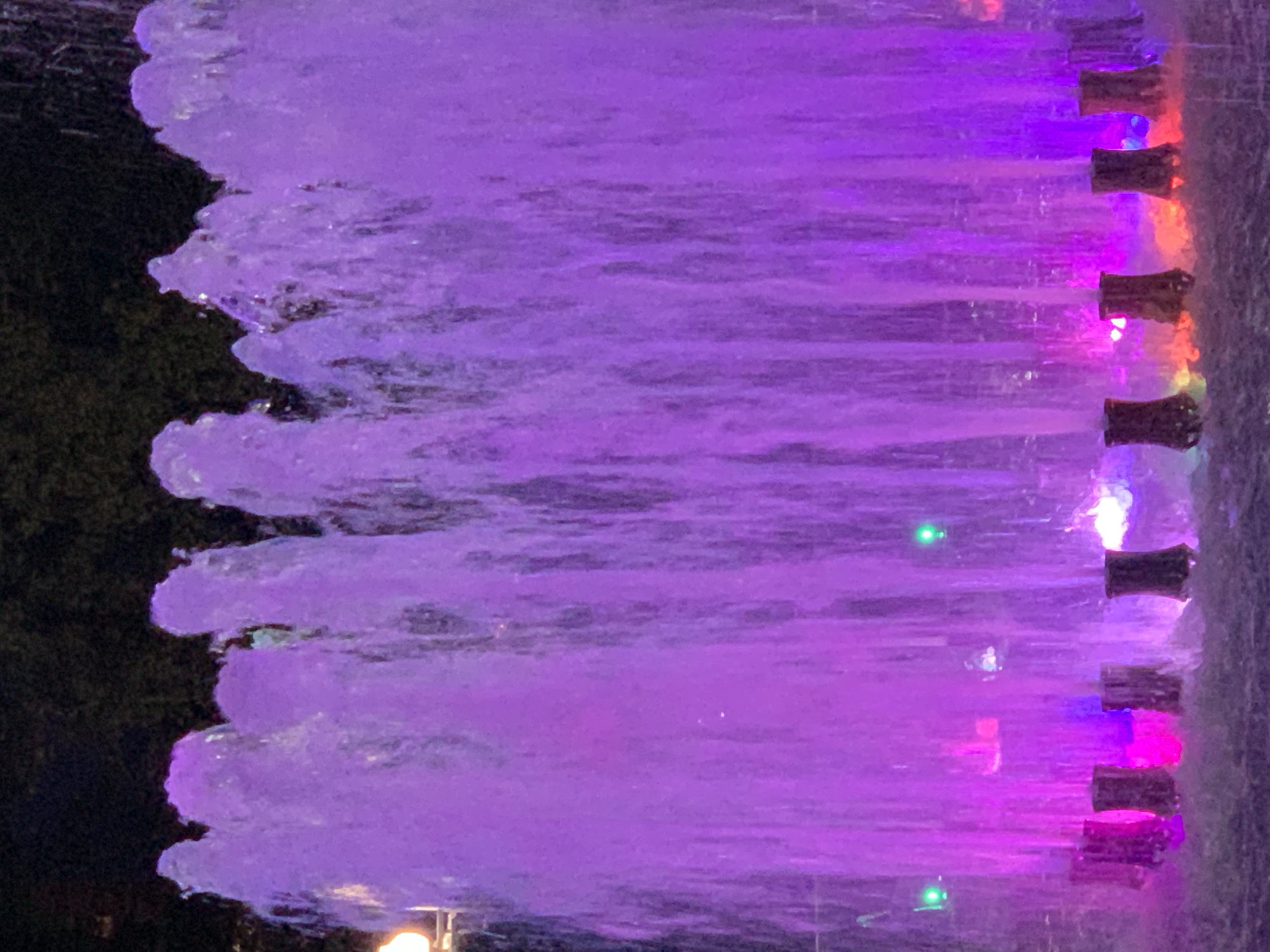 A nighttime exterior shot captures a fountain illuminated with vibrant purple lights. The water sprays upward in a dense, curtain-like fashion, creating a translucent effect as it cascades down. The base of the fountain is lined with several cylindrical structures, each emitting a different colored light, including pink, yellow, and red, which reflect off the water. Two small green lights are visible in the mid-ground of the fountain. The background is dark, suggesting it is nighttime, with faint foliage visible at the very top of the frame. The overall scene is one of a decorative outdoor fountain, likely a public display or park feature, at night.