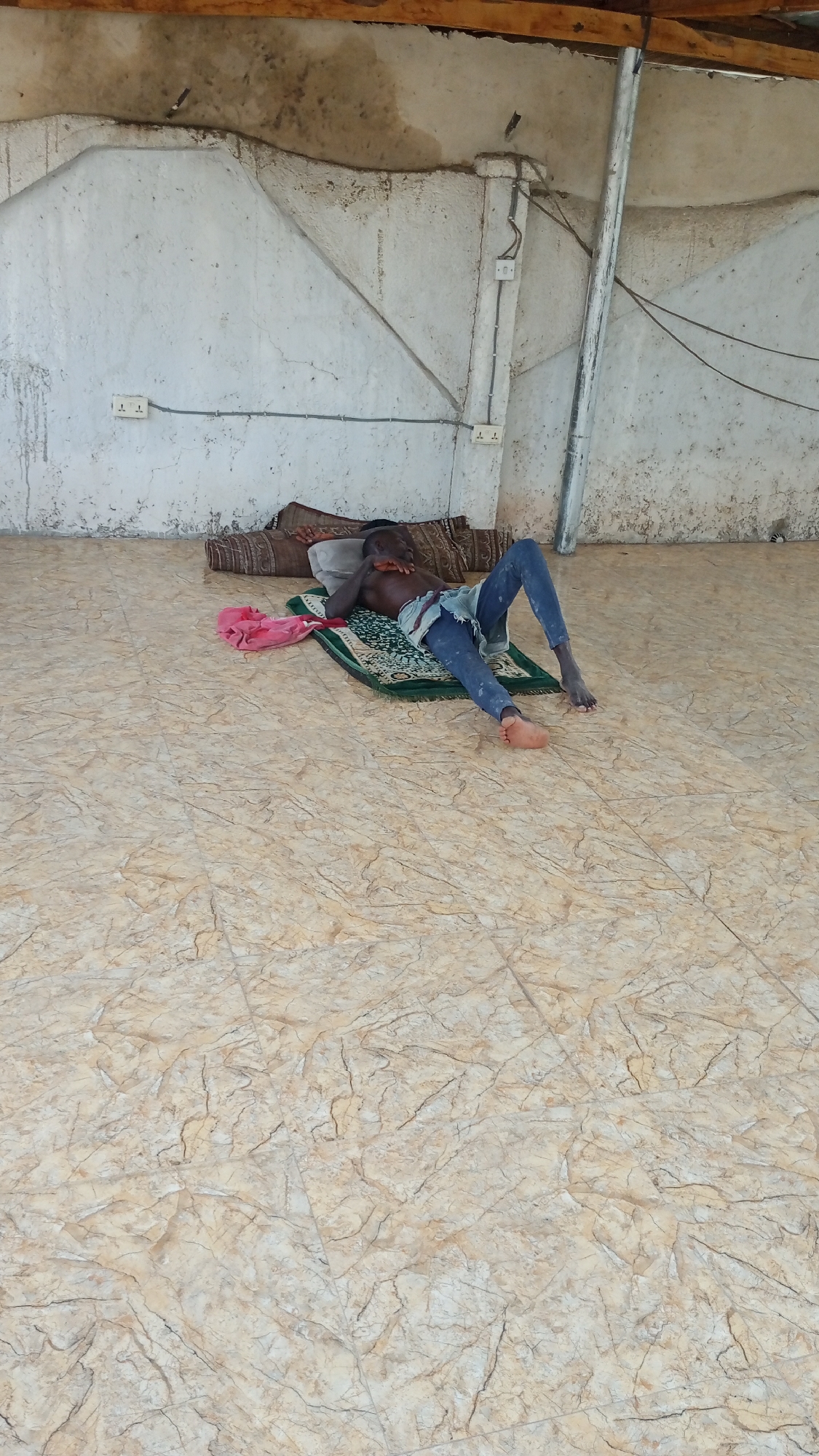 The image displays a young male, possibly a boy or young man, resting on the floor in what appears to be a simple, utilitarian indoor or semi-enclosed space in Lokoja, Nigeria. He is lying on his back, shirtless and clad in blue jeans, with his right arm bent and his hand partially obscuring his face. He is positioned on a green patterned mat, utilizing a rolled-up brown patterned rug as a pillow, with another similar rug behind his head. A pink fabric object, possibly a bag or garment, is visible near his left hip.
The setting is characterized by rough, light-colored walls exhibiting signs of wear, cracks, and discoloration. Exposed electrical wiring runs along the walls, connecting to two visible double electrical outlets marked "T T", one on the left and one on the right. A slender, light-colored vertical support beam is present on the right side of the frame. Overhead, rough wooden beams and an unfinished ceiling structure are visible. The floor is covered with light brown and beige tiles or linoleum, featuring a marbled or cracked pattern. The overall environment suggests a basic dwelling or shelter, consistent with a warm climate, which aligns with the individual's attire. The time of day appears to be daytime, though no direct light source is visible to confirm. The activity depicted is simply resting or sleeping.