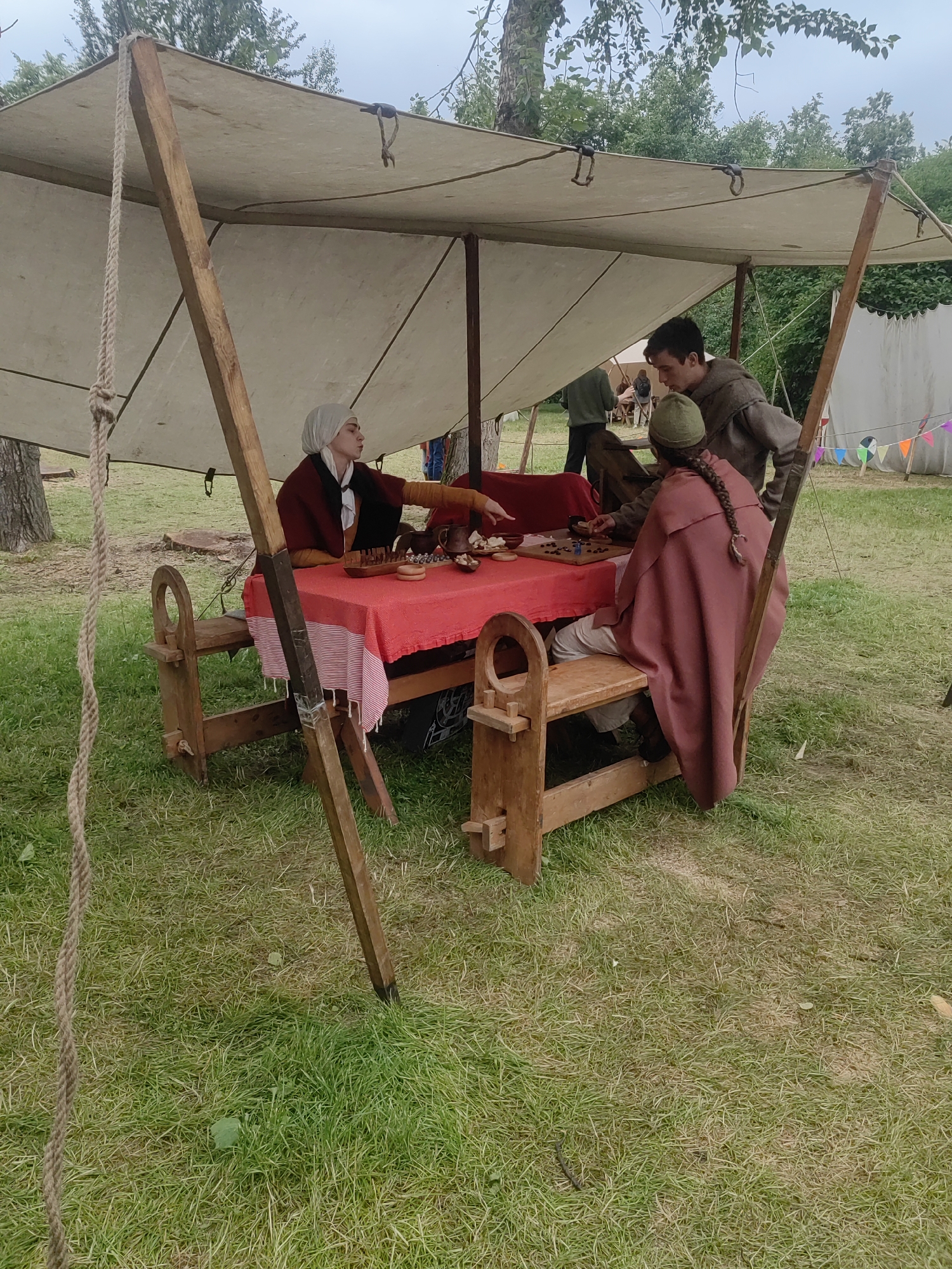 The image depicts a reenactment of a historical event, possibly a medieval fair or market, set outdoors on a grassy field. Three individuals are gathered around a red-covered table beneath a canvas tent supported by wooden poles.
On the left, a person wearing a light-colored headscarf, a dark red cloak over a yellow garment, and a white undershirt is seated, looking towards the table. They are gesturing with their right hand.
In the center, to the right of the first person, a person wearing a brown hooded tunic is leaning over the table, appearing to interact with the person on the far right.
On the far right, a person wearing a dark green hat and a reddish-pink cloak with a visible braid extending down their back is seated on a wooden bench. They are looking down at the table.
The table is set with various items, including what appears to be food or game pieces arranged on a wooden board, and a wooden bowl. The table itself has a red tablecloth and a striped cloth underneath. Wooden benches are positioned on either side of the table.
In the background, trees and more canvas tents are visible, suggesting a larger gathering. Decorative flags are strung between poles in the distance. The ground is covered in grass, with some patches of dry grass and fallen leaves. The sky is overcast.
There is no indication that this scene is located in Moscow, Russia.