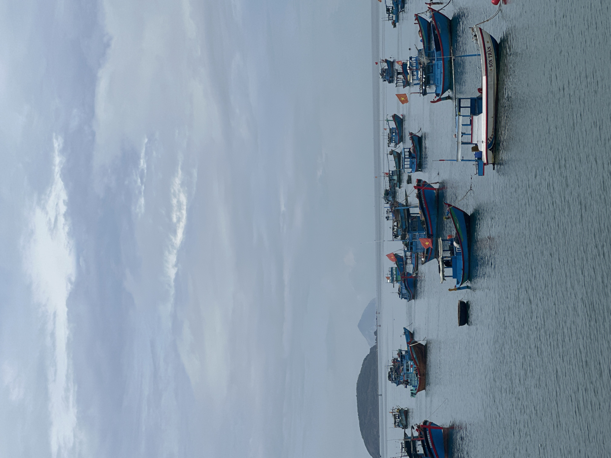The image depicts a harbor in Bac Nha Trang Ward, Vietnam, filled with numerous small fishing boats. Most are painted in blue hues with some red accents. Several have Vietnamese flags attached. In the background, land masses rise, appearing as hills or small mountains. The sky is overcast with a range of gray clouds. The water is calm and reflects the sky's somber tone.