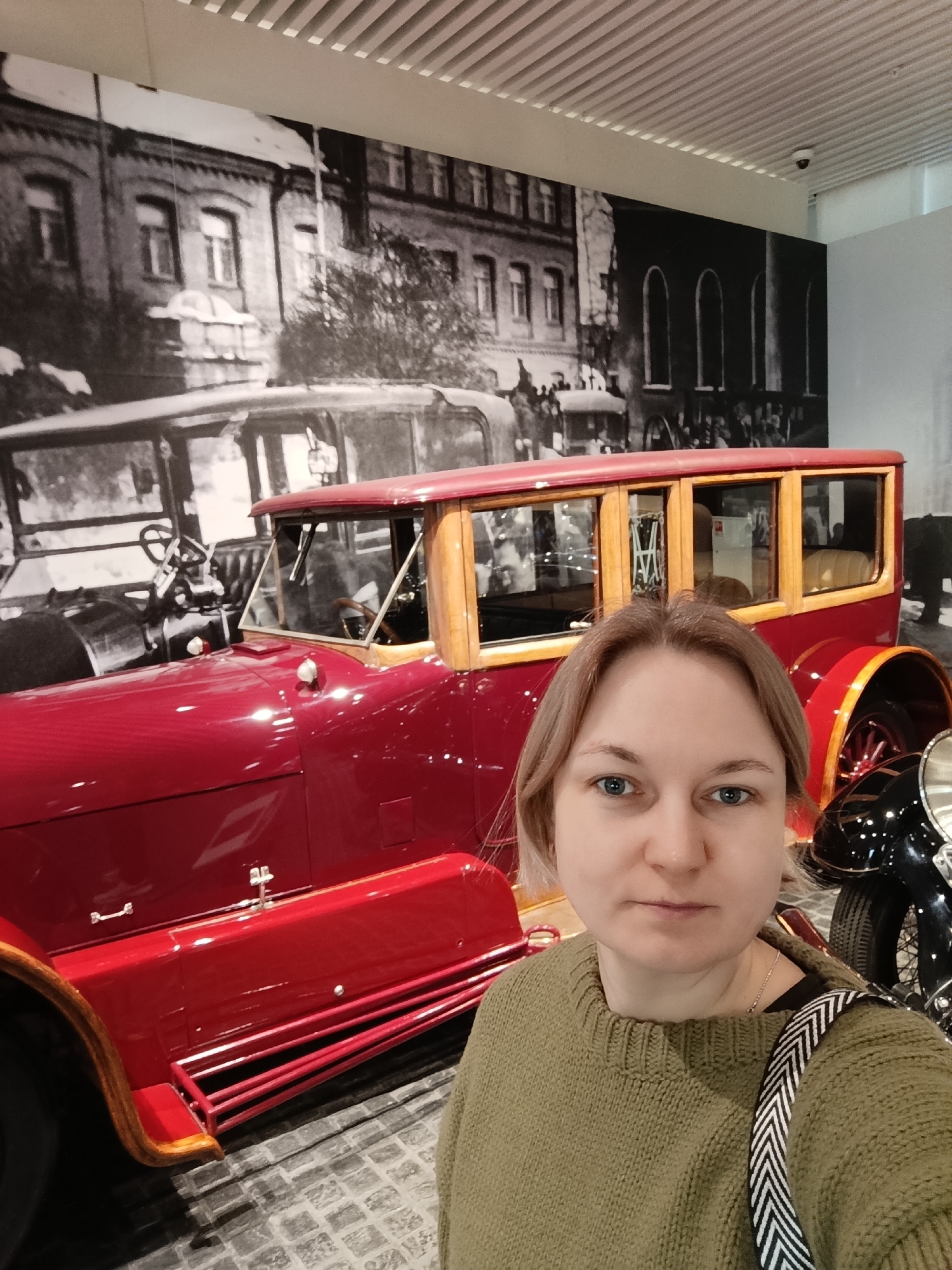 A woman with short blonde hair and blue eyes, wearing a green sweater and a black-and-white striped bag, is posing for a selfie in front of a vintage red car. The car has wooden trim and a classic design. Behind the car is a large black-and-white photograph of a building. The scene appears to be indoors, likely in a museum or exhibition space with a patterned floor. The woman is looking directly at the camera. This is most likely at the Ural Mining and Metallurgical Company's Museum of Military Equipment in Verkhnyaya Pyshma, Russia.
