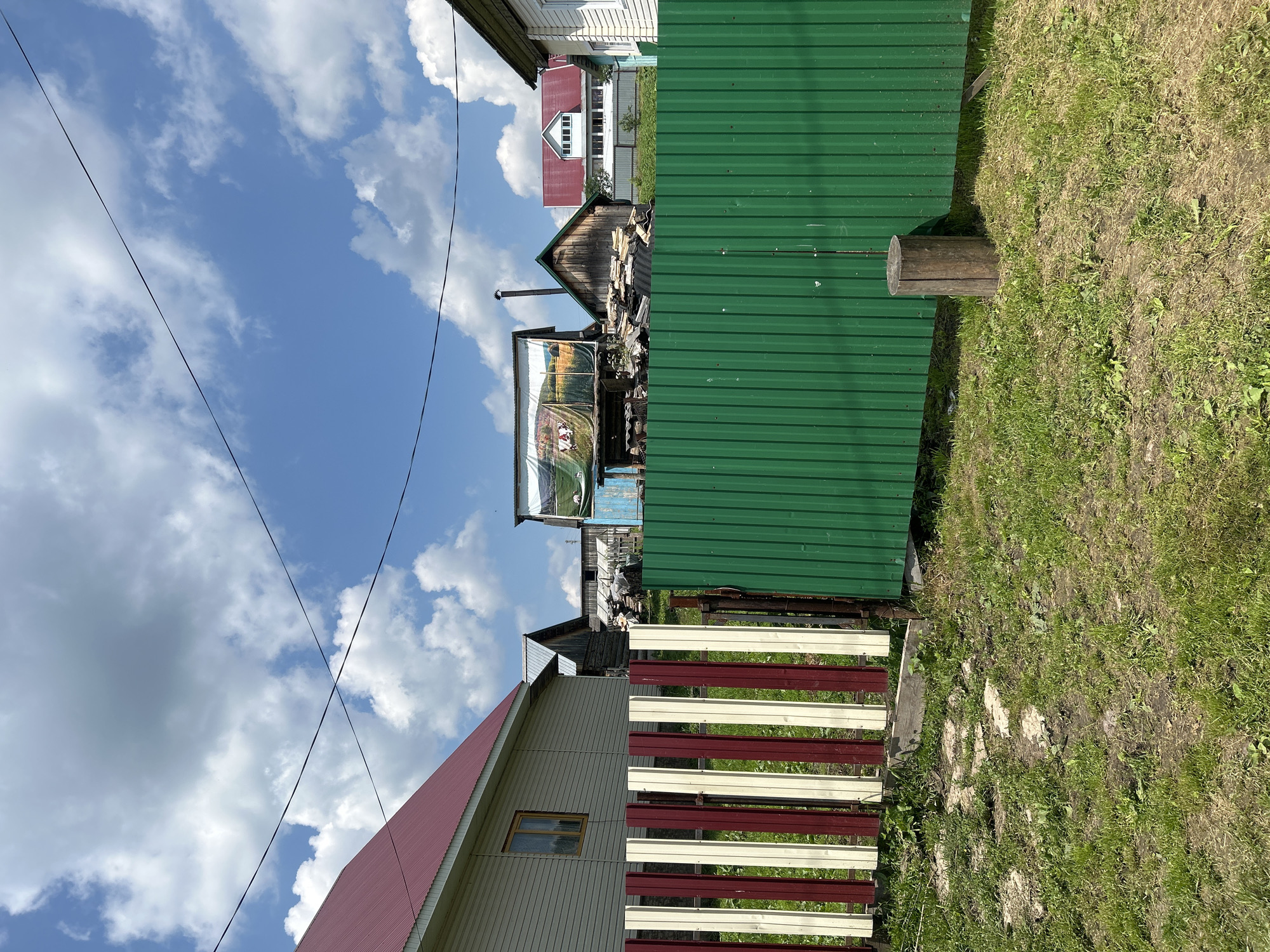 The image depicts a rural scene in Novomullakaevo, Russia, under a bright blue sky with scattered white clouds. In the foreground, a striped fence made of alternating red and cream vertical wooden planks is visible on the left, partially obscured by a green corrugated metal fence on the right. A wooden log stands upright in front of the green fence. Behind the fences, several structures are present. To the left, a light-colored house with a red metal roof and a single window is seen. Further back, various wooden buildings with different roof styles and materials are scattered across the landscape. One structure has a large banner or tarp displaying an image of cows in a field. The ground is covered in patchy green grass and dirt. Power lines are visible stretching across the sky.