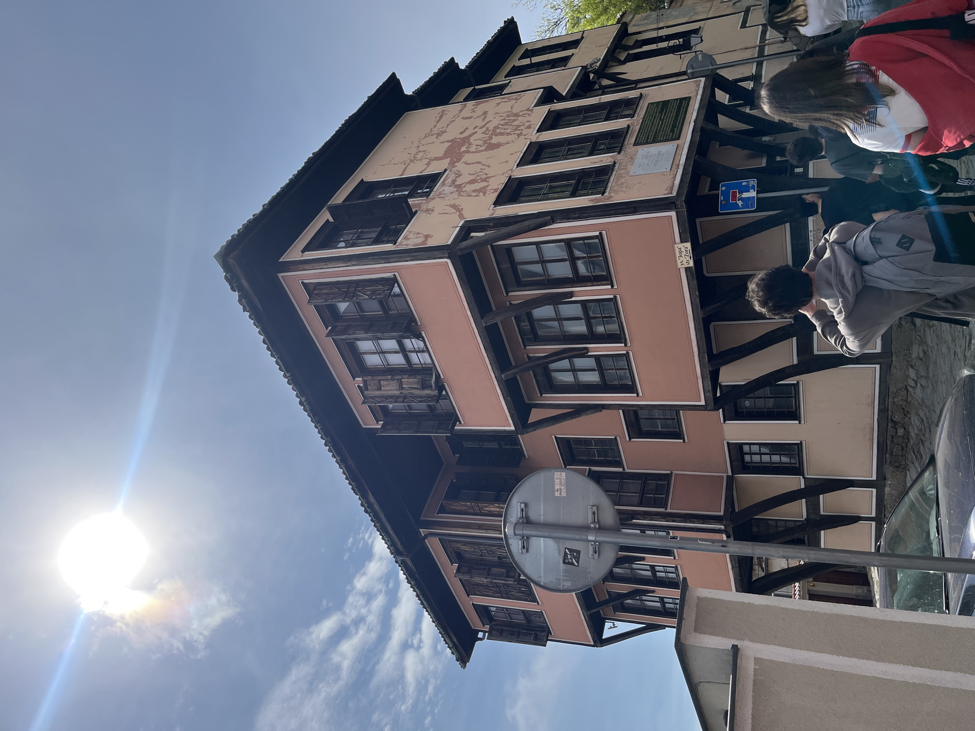 An upward-angled photograph captures a historic building on a bright, sunny day, likely in Plovdiv, Bulgaria, given the architectural style. The building features multiple stories with traditional Bulgarian National Revival architecture, characterized by its timber-framed structure and protruding upper floors, often called bay windows. The facade is painted in shades of peach, beige, and dark brown, with areas of faded and peeling paint on the upper levels. Numerous rectangular windows with dark frames are visible across all floors.
Several people are on the street adjacent to the building. One individual with a grey backpack is visible from behind in the foreground, seemingly looking at the building or an object nearby. Further back, another person in a red jacket and white top is partially visible, along with other individuals. They appear to be tourists or pedestrians exploring the area.
Notable details include the bright sun, which is prominently featured in the upper left quadrant of the upright image, creating a strong lens flare and glare. The sky is clear blue with a few scattered white clouds, indicating pleasant weather. A round, blue traffic sign with a white horizontal bar (typically indicating "no entry" or restricted access) is affixed to the building's facade. Below it, a smaller white rectangular sign bears text that reads "ул. Зорa" (ulitsa Zora), identifying the street as Zora Street. Two light-colored plaques are also mounted on the building's facade, likely historical markers, though their text is not clearly legible. Part of a dark car is visible parked on the street to the right. A large, grey circular sign with a metal pole, possibly another traffic sign, is seen from its back near the lower right of the frame. The overall impression is that of a bustling street scene in a historic European city.