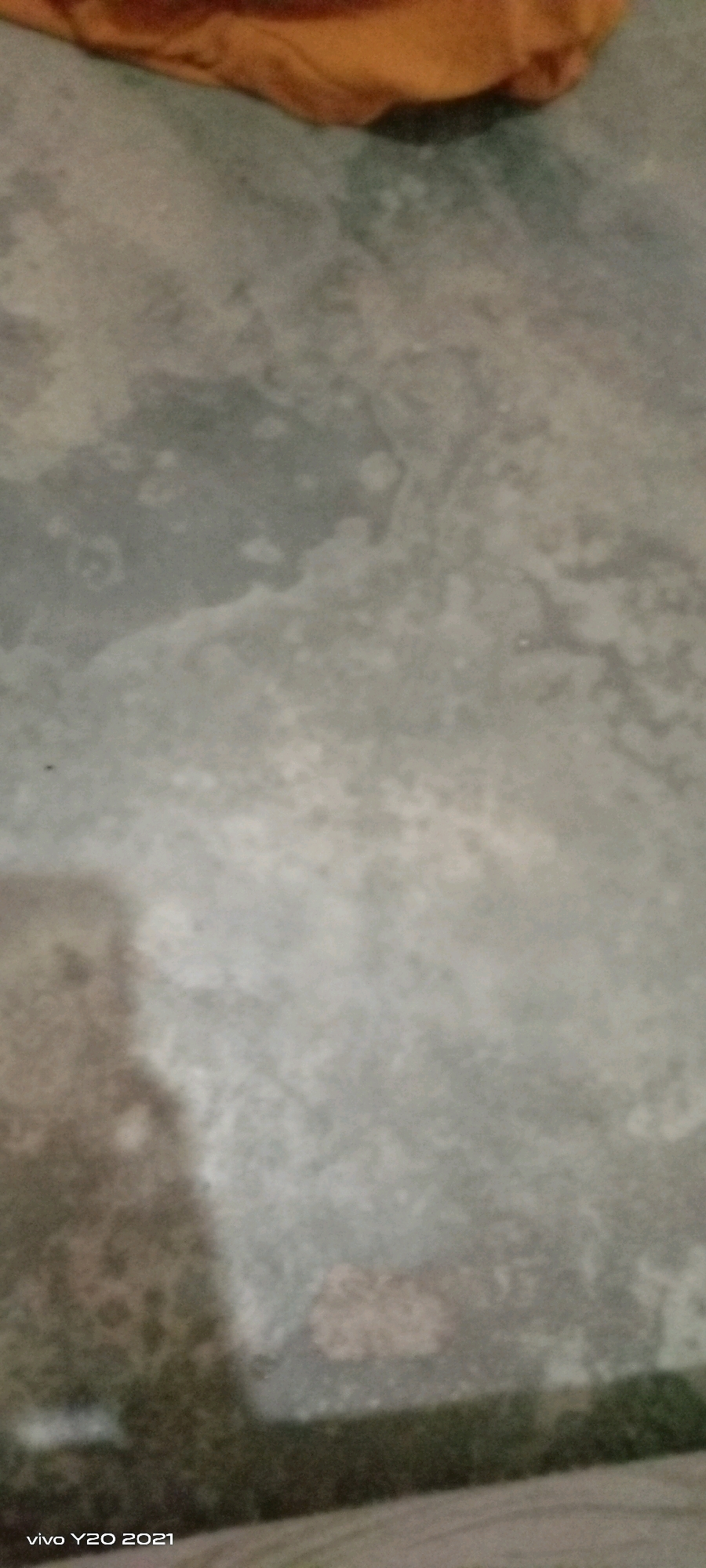 The image shows a close-up, slightly blurry view of a textured surface, possibly concrete or a similar material, with a darker shadow on the left side. In the upper portion of the image, a blurry object with an orange hue is visible. The bottom left corner of the image contains the text "vivo Y20 2021". The scene appears to be an indoor or outdoor ground surface. No specific location within a city or country can be determined from the visual information provided.
