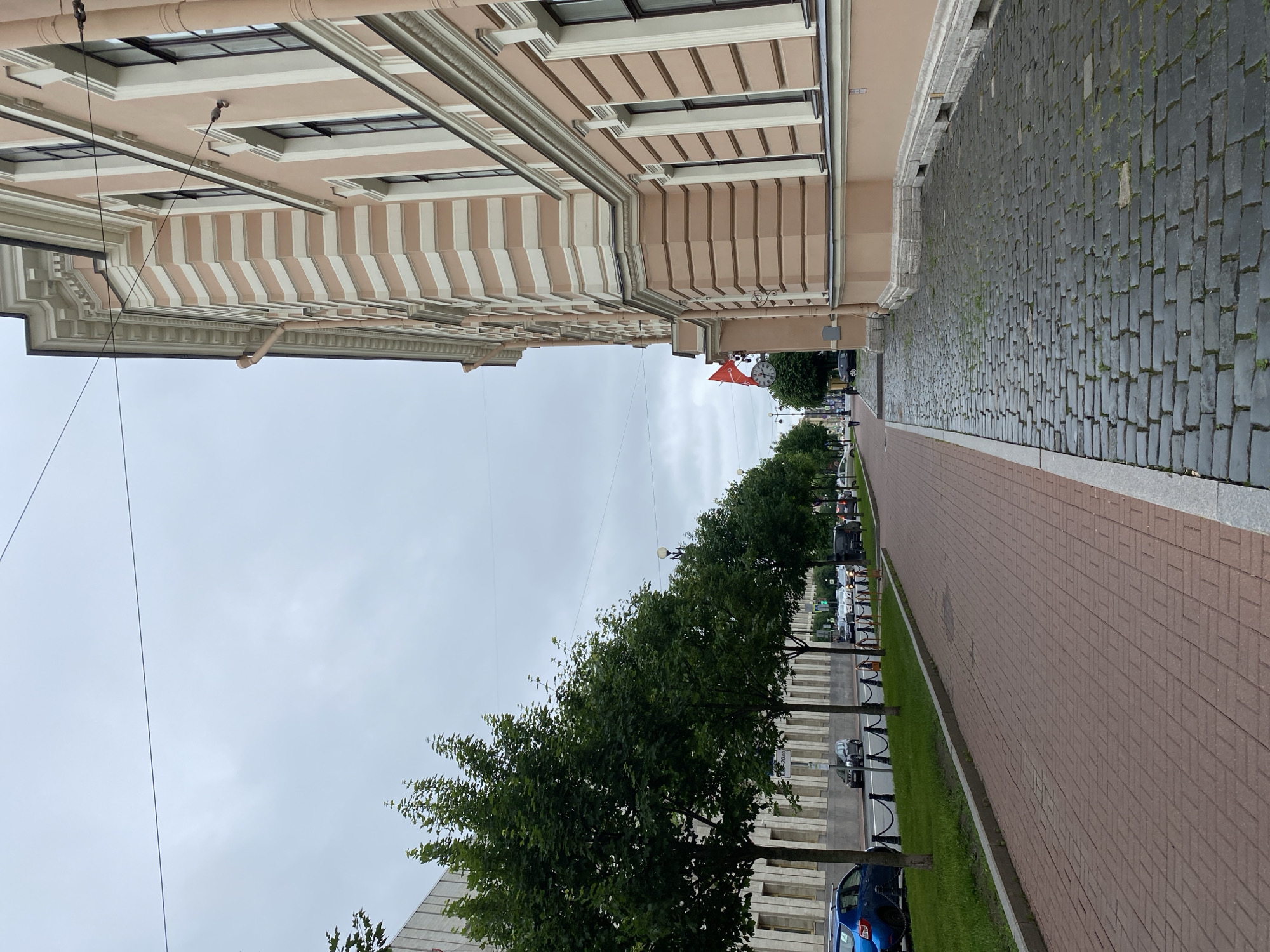 The image depicts a street scene in Saint Petersburg, Russia, with a large, ornate building on the right side and a tree-lined sidewalk and street on the left. The building is painted a pale pink with white decorative elements along its facade and features multiple windows. A clock and a red and white flag are visible on the side of the building in the middle ground. The sidewalk is paved with reddish bricks, and beyond that is a green lawn with a row of mature trees. The street is paved with dark cobblestones and has several cars parked and moving. The sky is overcast.