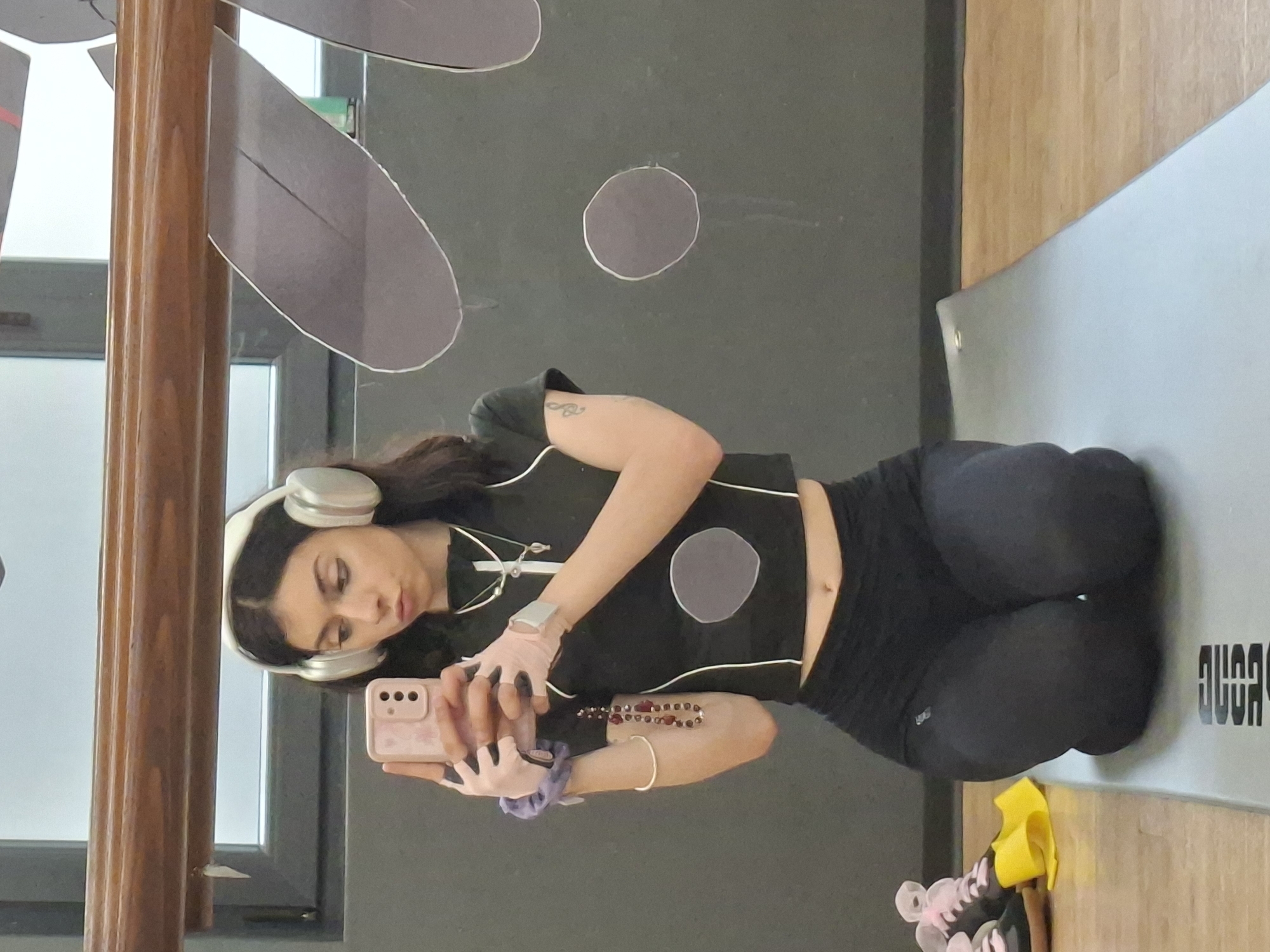 The image, oriented horizontally but depicting a standing figure, shows a young woman taking a mirror selfie in what appears to be an indoor fitness environment, possibly a gym or studio in Sofia, Bulgaria. She has long dark hair and is wearing white over-ear headphones, a black short-sleeved crop top with white trim and a front zipper/clasp, and black high-waisted athletic leggings.
Her hands are adorned with light pink fingerless gloves, and she wears several bracelets, including a red beaded one and a silver bangle, with a purple scrunchie on her left wrist. She is holding a light pink smartphone with a matching case, looking down at the screen. A small tattoo is visible on her upper right arm, and a subtle belly button piercing can be seen.
The background includes a dark wall decorated with abstract grey circular and elongated shapes outlined in white. A wooden column with vertical grooves is visible on the left, and a wooden floor on the right. She is standing on a grey exercise mat, where partially visible, mirrored text resembling "AGOY OLA" (likely a reversed brand name such as "ALO YOGA") can be seen. On the floor nearby are pink and black athletic shoes and a yellow resistance band. The scene suggests she is taking a break during or before a workout session.