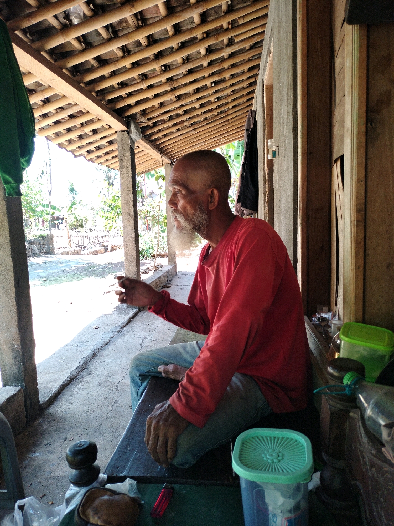 The image depicts a man with a shaved head and a beard sitting on a wooden bench under a bamboo-thatched roof. He is wearing a red long-sleeved shirt and blue jeans. The man is holding a cigarette in his right hand, which appears to be lit. In the foreground, there are several objects on the bench, including two black, stacked items that resemble weights, a small brown pouch, and a red lighter. To the right, there are two green plastic containers, one with a lid. The environment suggests a rural or semi-rural setting, with a concrete floor and the suggestion of outdoor vegetation visible in the background. The architecture, with its exposed bamboo roof structure and wooden walls, is consistent with common building styles in rural Indonesia, including the area of Karangmojo. The lighting indicates it is daytime, with bright sunlight casting shadows.