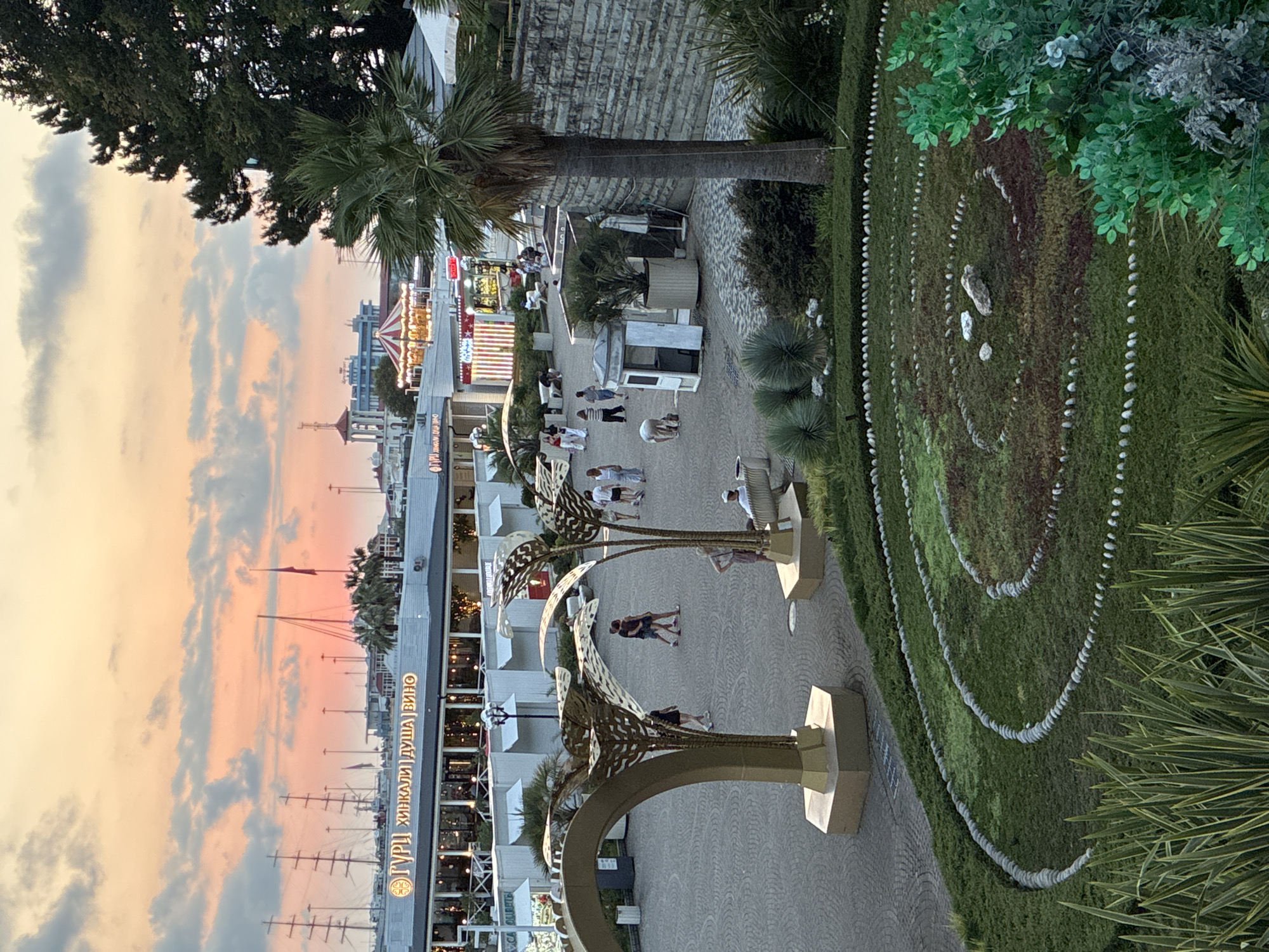 The image depicts an outdoor scene in Sochi, Russia, likely during sunset. In the foreground, a circular garden bed is visible, featuring concentric circles of stones and various green and reddish-brown ground cover. Tall, spiky green plants line the bottom edge of the frame.
In the middle ground, a paved plaza area is populated by several individuals. Some are walking, while others are seated or interacting. Two people are walking together towards the left, and a group of four is visible near the center, heading towards the right. A lone figure is seated on a bench to the right of the plaza. Several abstract golden sculptures, resembling palm leaves, are positioned within the plaza. A small, white booth-like structure stands near the center.
The background reveals a row of buildings and stalls, some illuminated with decorative lights. A carousel with a red and white striped roof is prominent in the distance, with lights on. Beyond the buildings, tall sailing ships with masts are docked in a harbor. The sky is filled with dramatic clouds, tinged with orange and pink hues, suggesting the late afternoon or early evening. A large, dark green tree dominates the right side of the frame, partially obscuring the background. The overall atmosphere is one of a bustling waterfront area at dusk.
