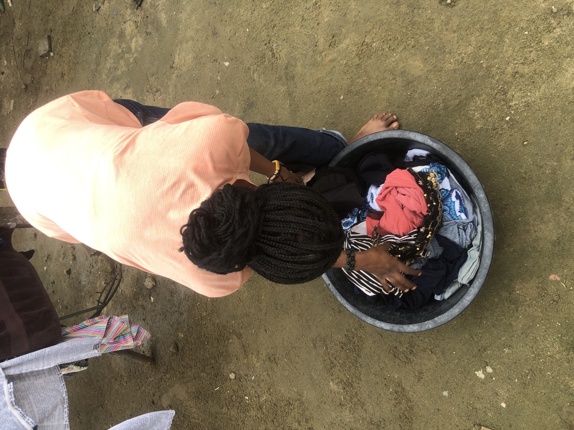 In Ado, Nigeria, a person wearing a light peach-colored, short-sleeved shirt and dark blue jeans is bent over a large, black plastic basin filled with laundry. The person has dark hair styled in intricate braids pulled up into a bun. Their right hand is reaching into the basin, sorting through the clothes, which include striped, patterned, and solid-colored garments. A bracelet with yellow beads adorns their wrist. The ground around the basin is composed of dirt and small stones. In the background, a clothesline with various fabrics is visible, supported by metal poles. The overall setting appears to be an outdoor, possibly domestic, area.