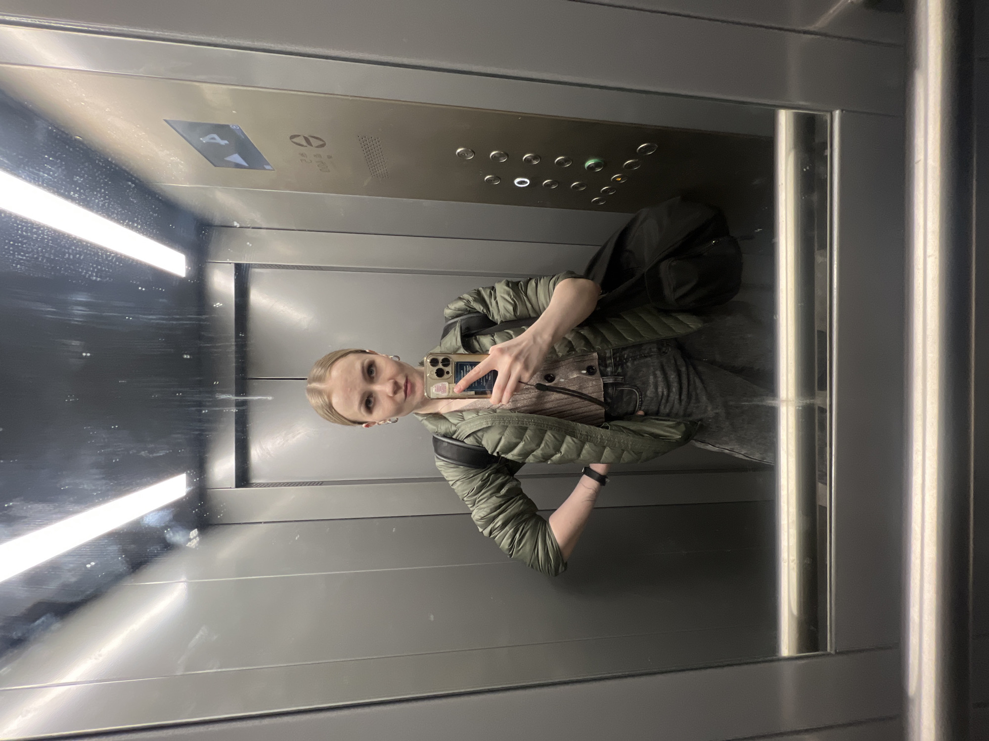 A woman takes a selfie in an elevator. The elevator has a metal interior. The woman wears a green puffer jacket and a black backpack. The elevator buttons are visible. The elevator is labeled with the number 4. The location is Khimki, Russia.