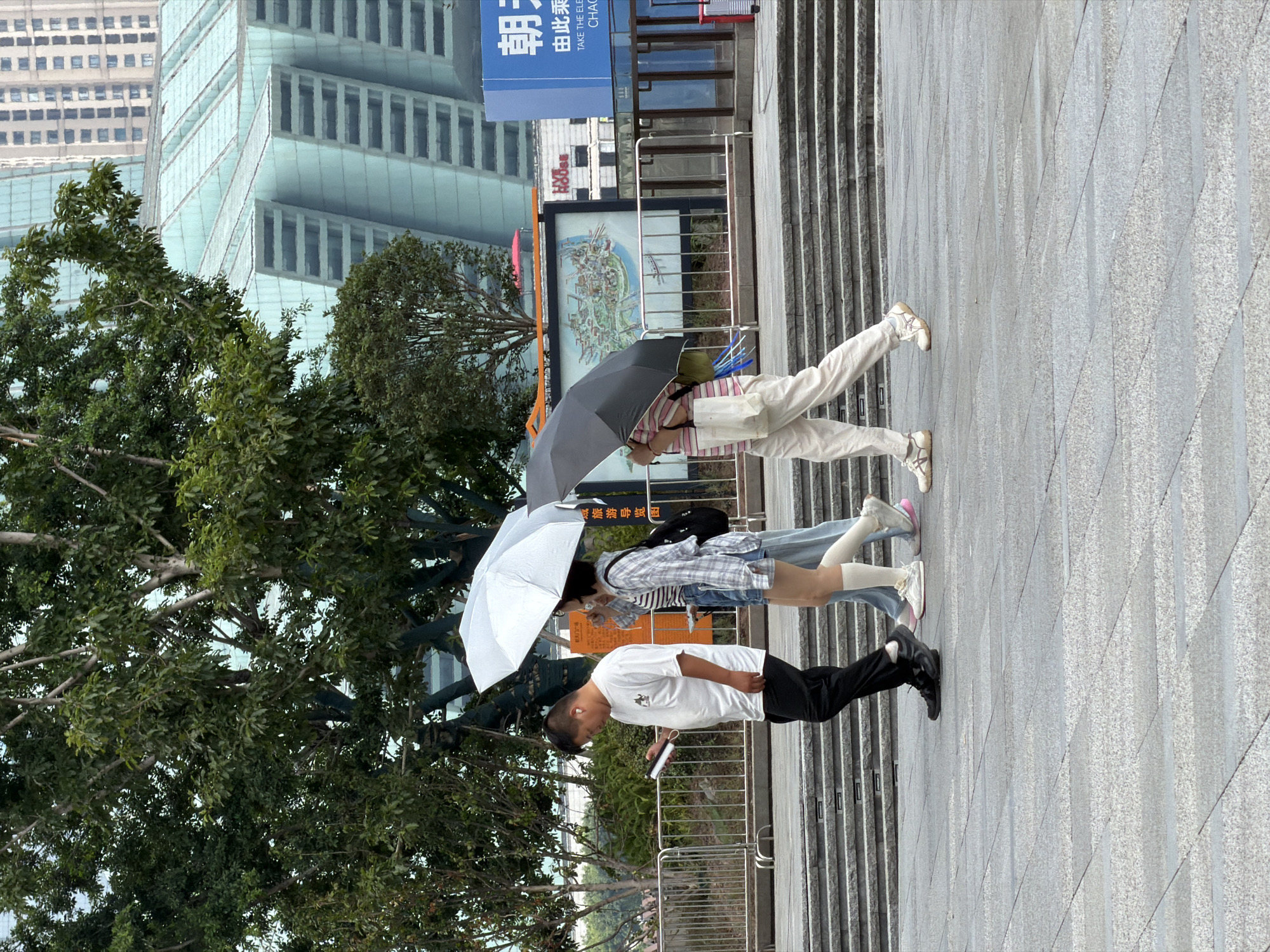 In the Yuzhong District of China, three people are walking on a paved outdoor area. The person on the left is a male wearing a white t-shirt, black pants, and black shoes, holding a smartphone and wearing wireless earbuds. In the middle, a female with short dark hair wears a checkered shirt, denim shorts, white knee-high socks, and white sneakers. She is holding a white umbrella over herself and the person to her right. The third person, on the right, is a female wearing a striped t-shirt, light-colored pants, and light-colored sneakers. She is holding a dark umbrella. Behind the individuals, a metal railing separates the walkway from a tiered stone staircase. Beyond the railing, trees are visible, and in the background, a large, modern building with a glass facade and a high-rise building are present. A blue sign with Chinese characters is also visible in the background. The ground appears slightly wet, suggesting recent rain.